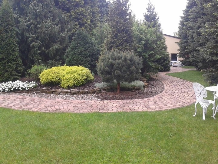 Ogród z krętą ścieżką z czerwonej kostki brukowej, otoczony zielonym trawnikiem, ozdobnymi krzewami i wysokimi drzewami iglastymi. Białe, metalowe krzesła i stolik w prawym dolnym rogu.