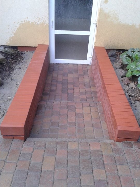 Ułożona kostka brukowa prowadząca do drzwi wejściowych, z ceglanymi obrzeżami po bokach, widoczne różnice w odcieniach kostki i niewielkie zabrudzenia na ścianie budynku.