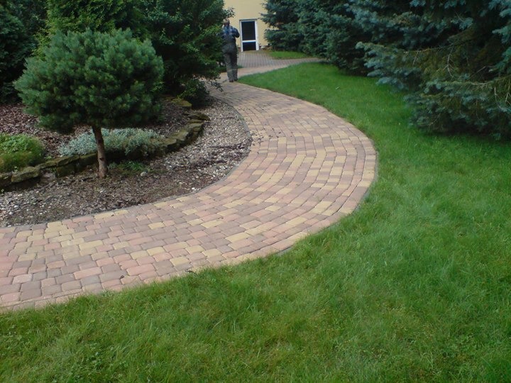 Zakrzywiony chodnik z kostki brukowej w odcieniach beżu i brązu, otoczony trawnikiem i rabatą z ozdobnymi krzewami, z sylwetką osoby w tle.