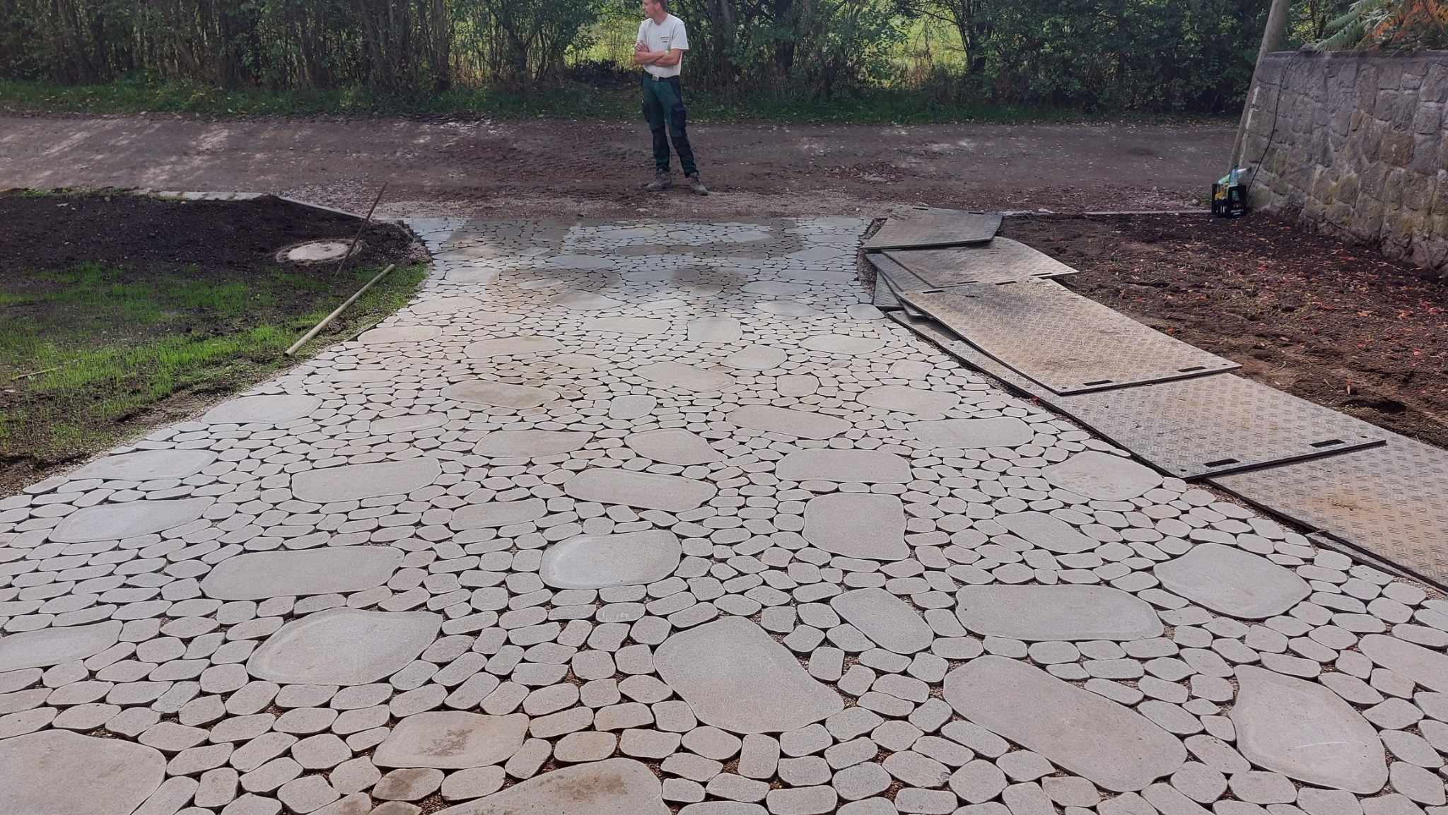 Ścieżka z nieregularnych, jasnoszarych kamieni ułożona w ogrodzie, widoczny pracownik w oddali i płyty ochronne na ziemi.