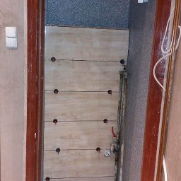 GLAREM - Montaż płytek imitujących drewno na ścianie, widoczne otwory na rury, system poziomowania płytek na podłodze, drzwi z ciemnego drewna, instalacja wodna i elektryczna w tle.