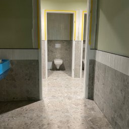 GLAREM - Wnętrze łazienki w trakcie prac wykończeniowych; widoczne płytki imitujące marmur na podłodze i ścianach, zabezpieczone taśmą malarską ościeżnice drzwi, w głębi toaleta z podtynkowym systemem...