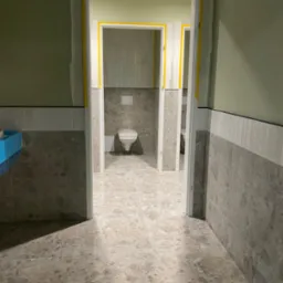 Wnętrze łazienki w trakcie prac wykończeniowych; widoczne płytki imitujące marmur na podłodze i ścianach, zabezpieczone taśmą malarską ościeżnice drzwi, w głębi toaleta z podtynkowym systemem...