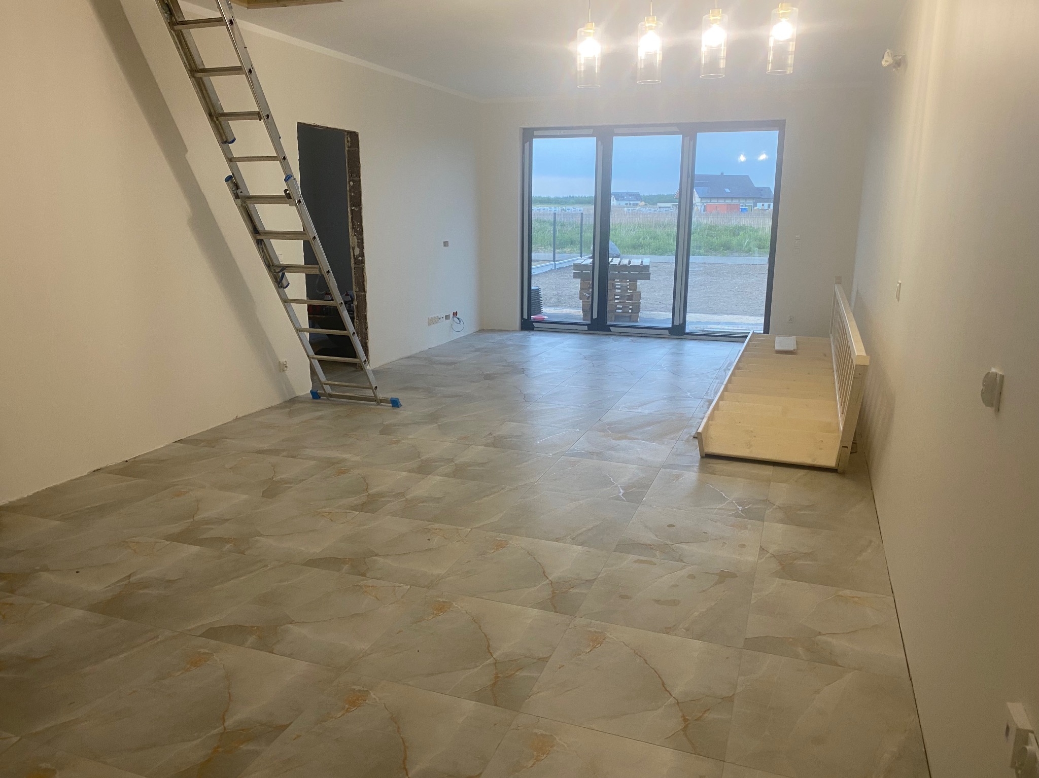 Wnętrze w stanie deweloperskim z marmurową podłogą, schodkami prowadzącymi do okna balkonowego i aluminiową drabiną opartą o ścianę, gotowe do prac wykończeniowych.