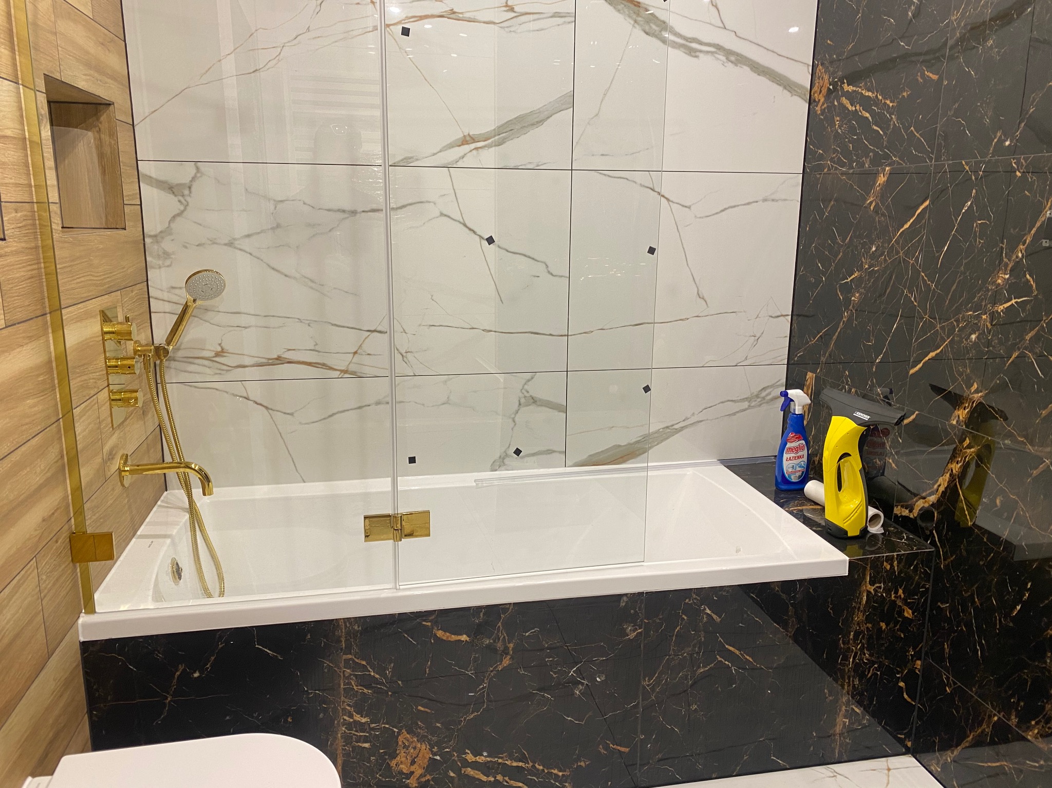 Nowoczesna łazienka z wanną obudowaną czarnymi płytkami imitującymi marmur, białe płytki marmurowe na ścianach, złota armatura prysznicowa i szklany parawan nawannowy. Widoczny środek...