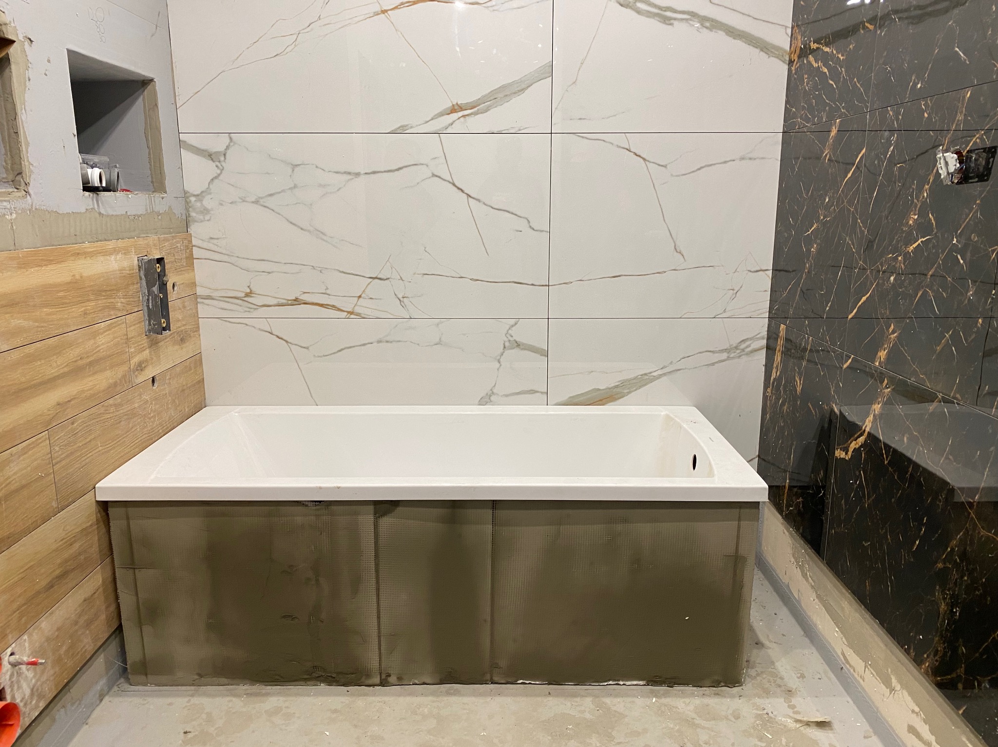 Montaż wanny w łazience z częściowo ułożonymi płytkami imitującymi marmur w kolorze białym i czarnym ze złotymi żyłkami. Obudowa wanny w trakcie wykańczania, zabezpieczona siatką i klejem.