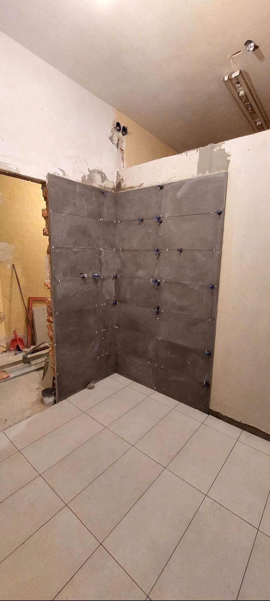 Świeżo ułożone, szare płytki ścienne w rogu łazienki, zabezpieczone niebieskimi krzyżykami dystansowymi, kontrastują z jasną podłogą z płytek i surowym wykończeniem ścian.