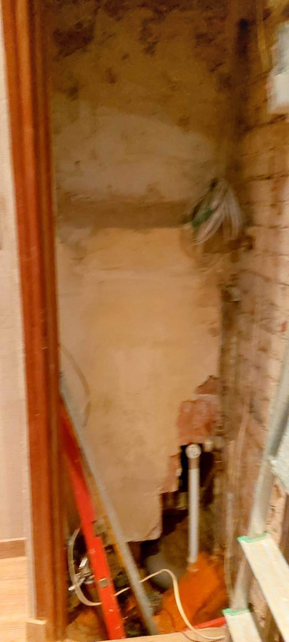 Niedokończona wnęka w łazience z odsłoniętymi rurami, przewodami elektrycznymi i fragmentami starego tynku na ceglanej ścianie, widoczne narzędzia budowlane i drabina.