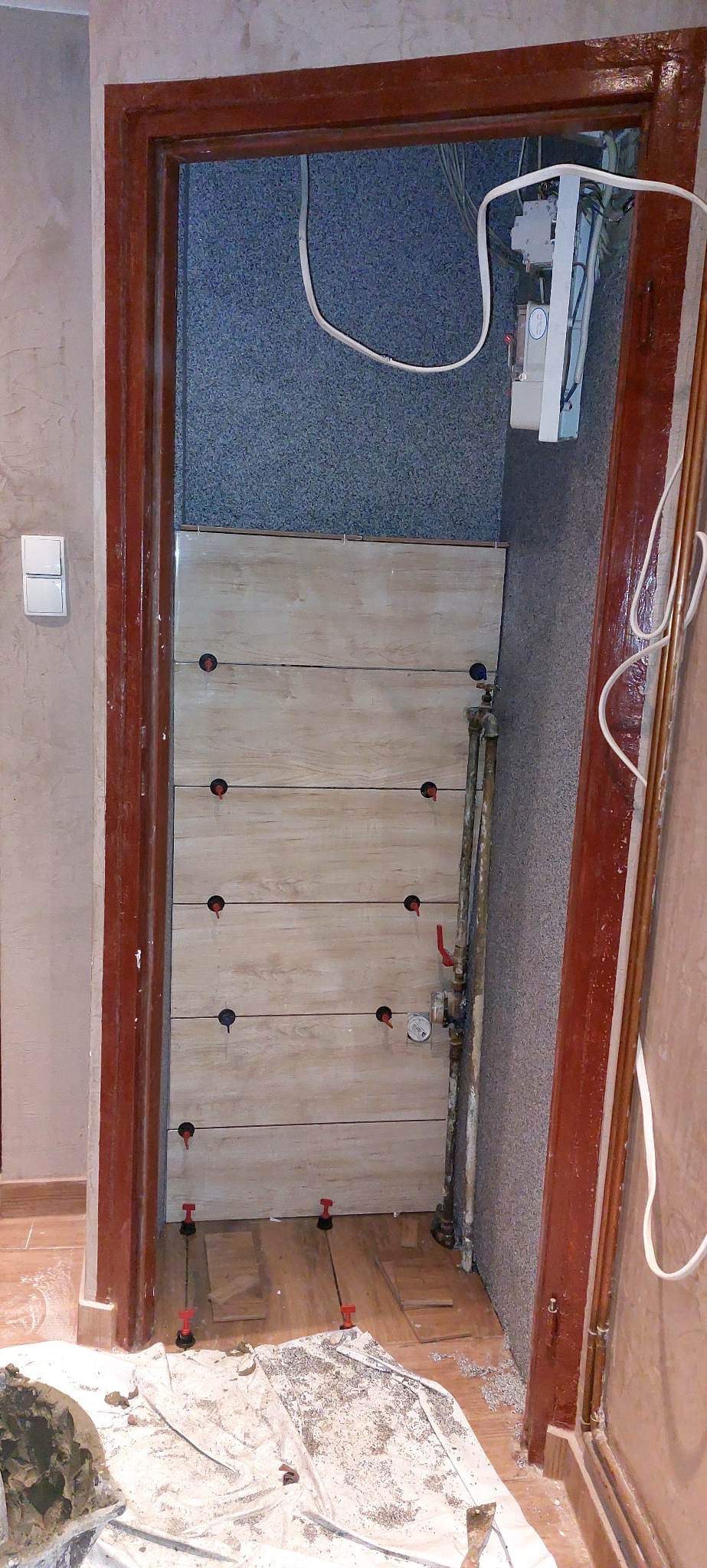 Montaż płytek imitujących drewno na ścianie, widoczne otwory na rury, system poziomowania płytek na podłodze, drzwi z ciemnego drewna, instalacja wodna i elektryczna w tle.