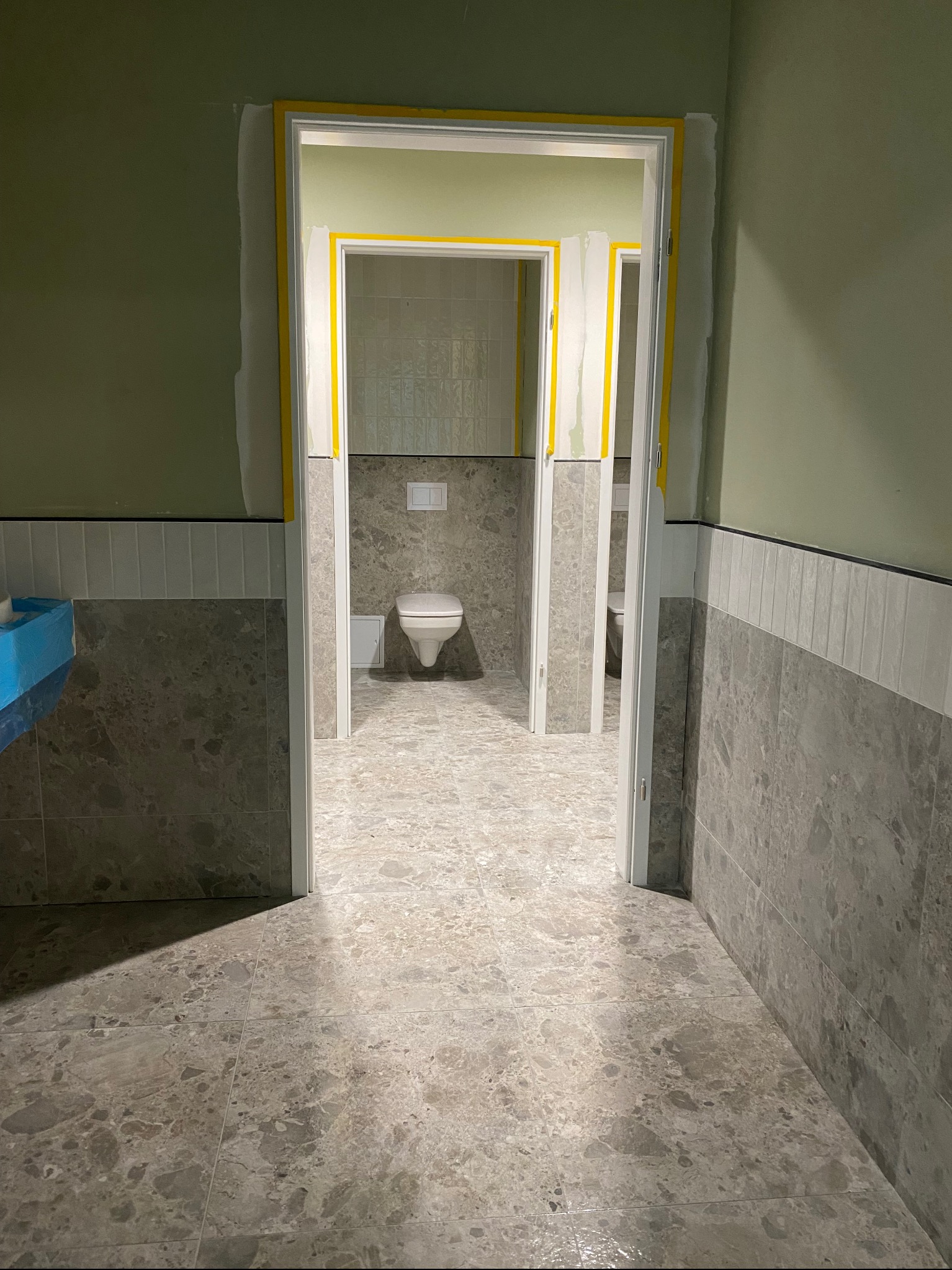 Wnętrze łazienki w trakcie prac wykończeniowych; widoczne płytki imitujące marmur na podłodze i ścianach, zabezpieczone taśmą malarską ościeżnice drzwi, w głębi toaleta z podtynkowym systemem...