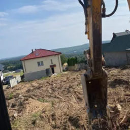 Żółta koparka z łyżką w trakcie niwelacji terenu pod budowę domu z czerwoną dachówką na wzgórzu, widoczne ślady gąsienic na ziemi.