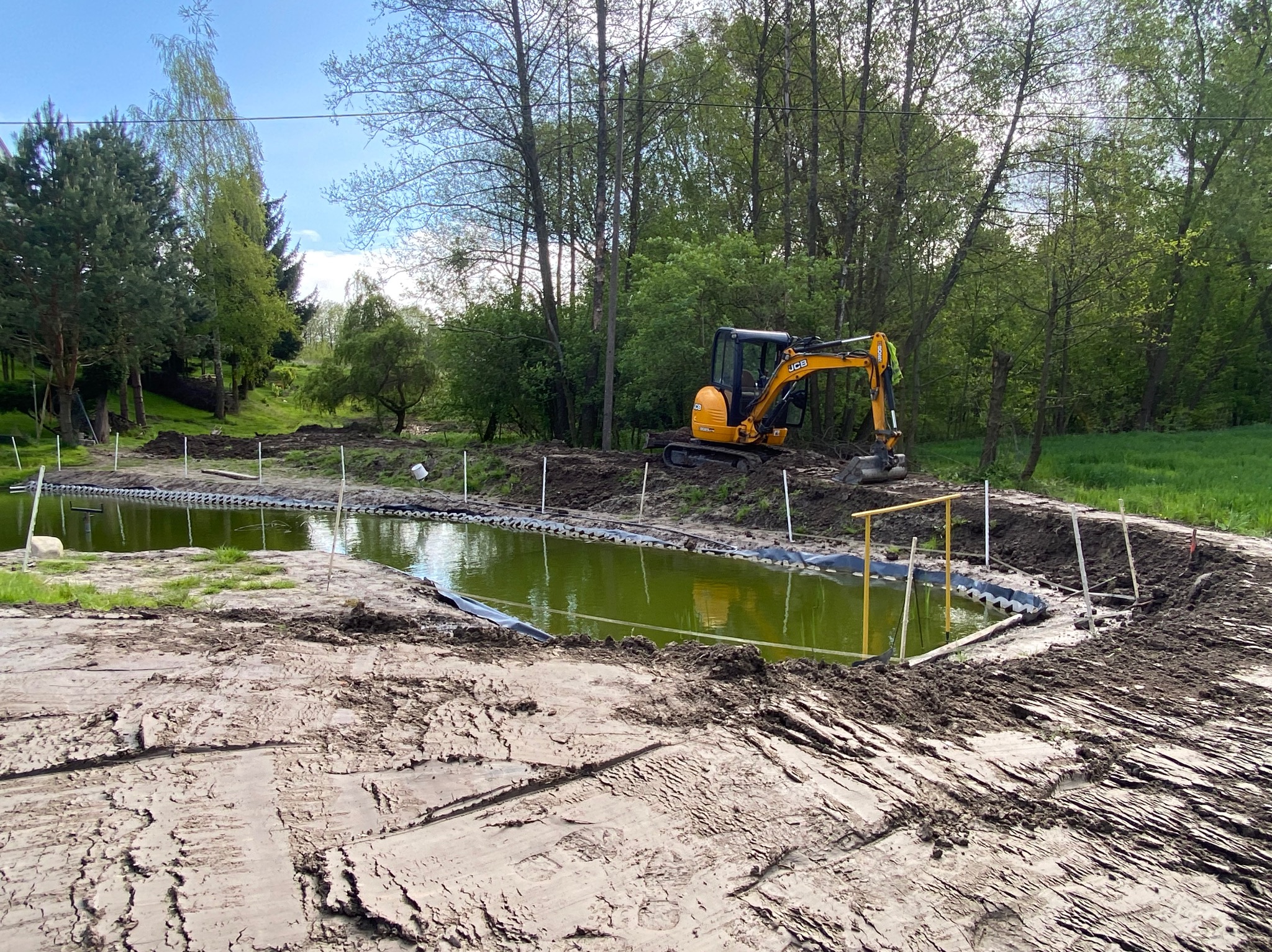 Żółta minikoparka JCB przy budowie oczka wodnego z wykopanym dołem wyłożonym czarną folią i wzmocnionym konstrukcją, w tle drzewa i trawa.