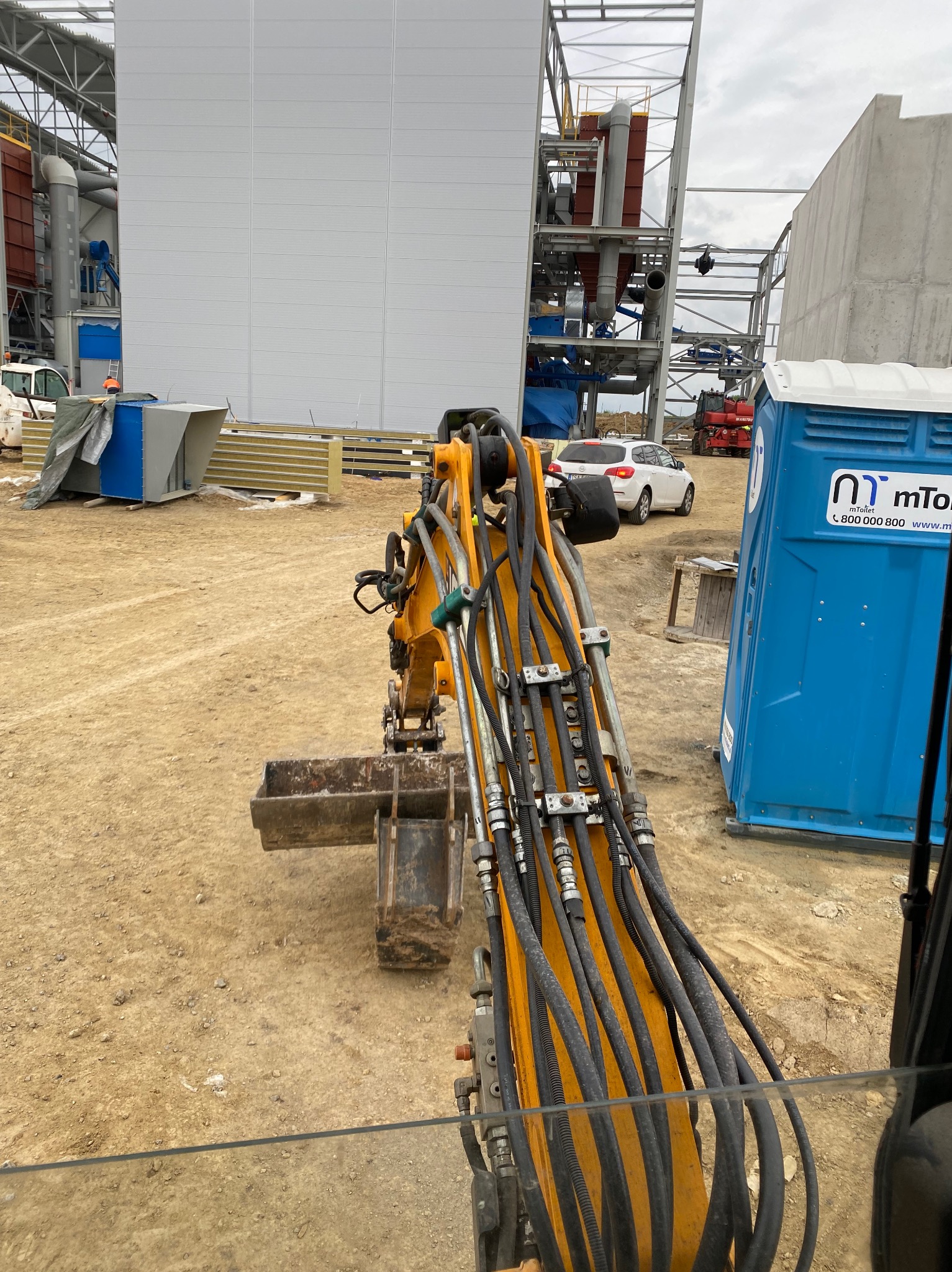 Widok z kabiny żółtej mini koparki na placu budowy z widocznymi przewodami hydraulicznymi, w tle hala przemysłowa i przenośna toaleta.