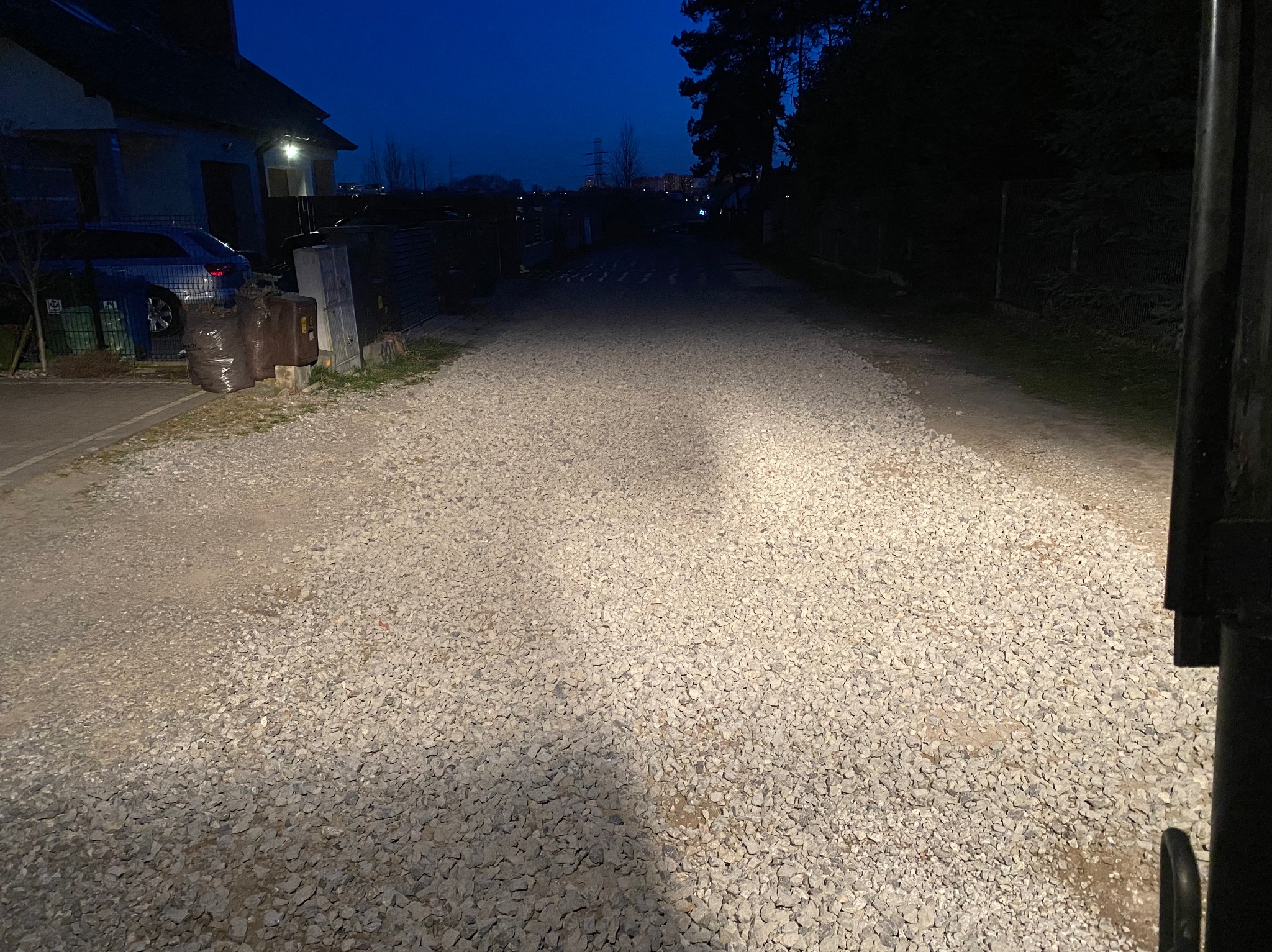 Utwardzona droga żwirowa w słabym oświetleniu, widoczne domy w tle i słupy wysokiego napięcia w oddali.