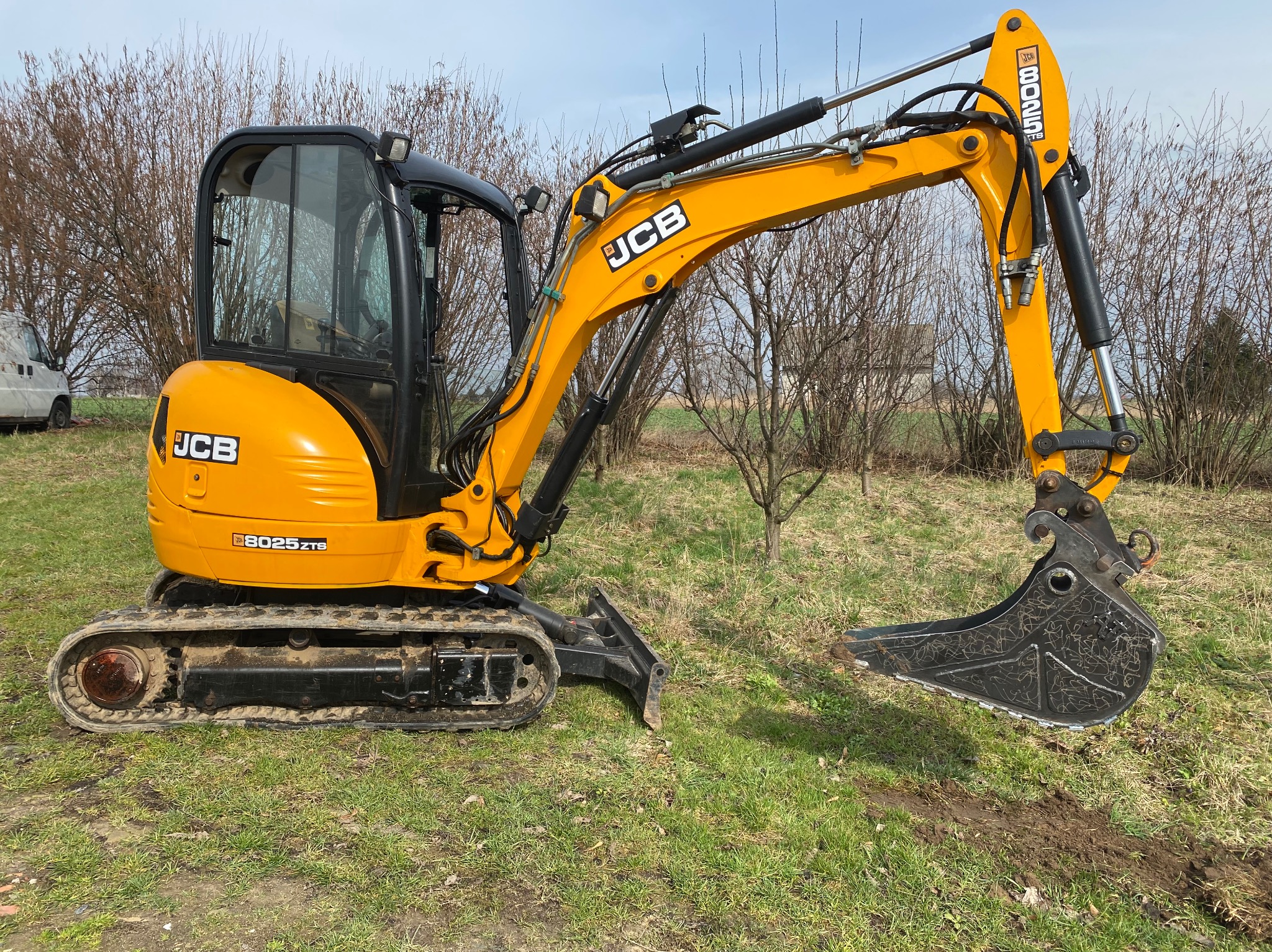 Żółta mini koparka JCB 8025 ZTB stoi na trawie z łyżką skierowaną w dół, w tle nagie krzewy i częściowo widoczny biały samochód dostawczy.
