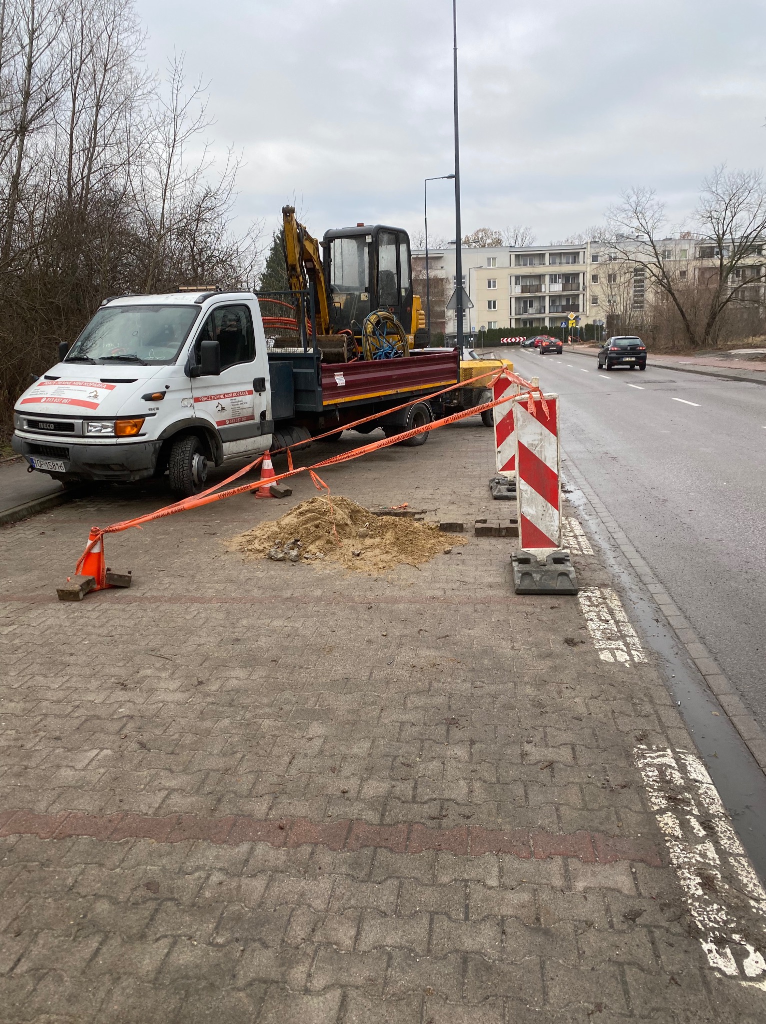 Mała koparka na platformie samochodu ciężarowego zaparkowana na chodniku obok wykopu zabezpieczonego taśmą ostrzegawczą i znakami drogowymi, w tle ulica i budynki mieszkalne.