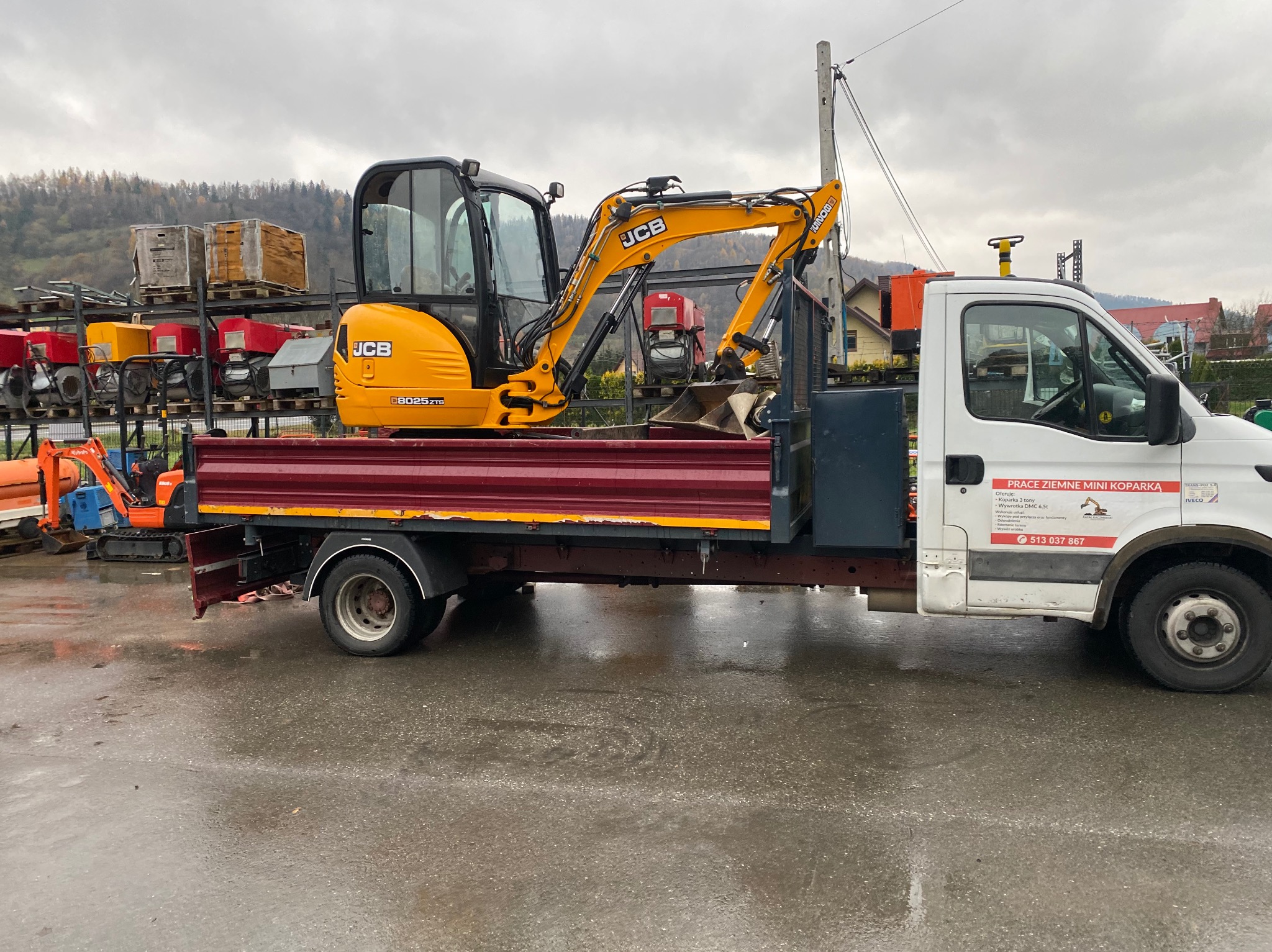 Żółta mini koparka JCB 8025ZTS na czerwonej platformie samochodu dostawczego z widocznym numerem telefonu i napisem 'PRACE ZIEMNE MINI KOPARKĄ' na boku, w tle inne maszyny budowlane i zalesione...