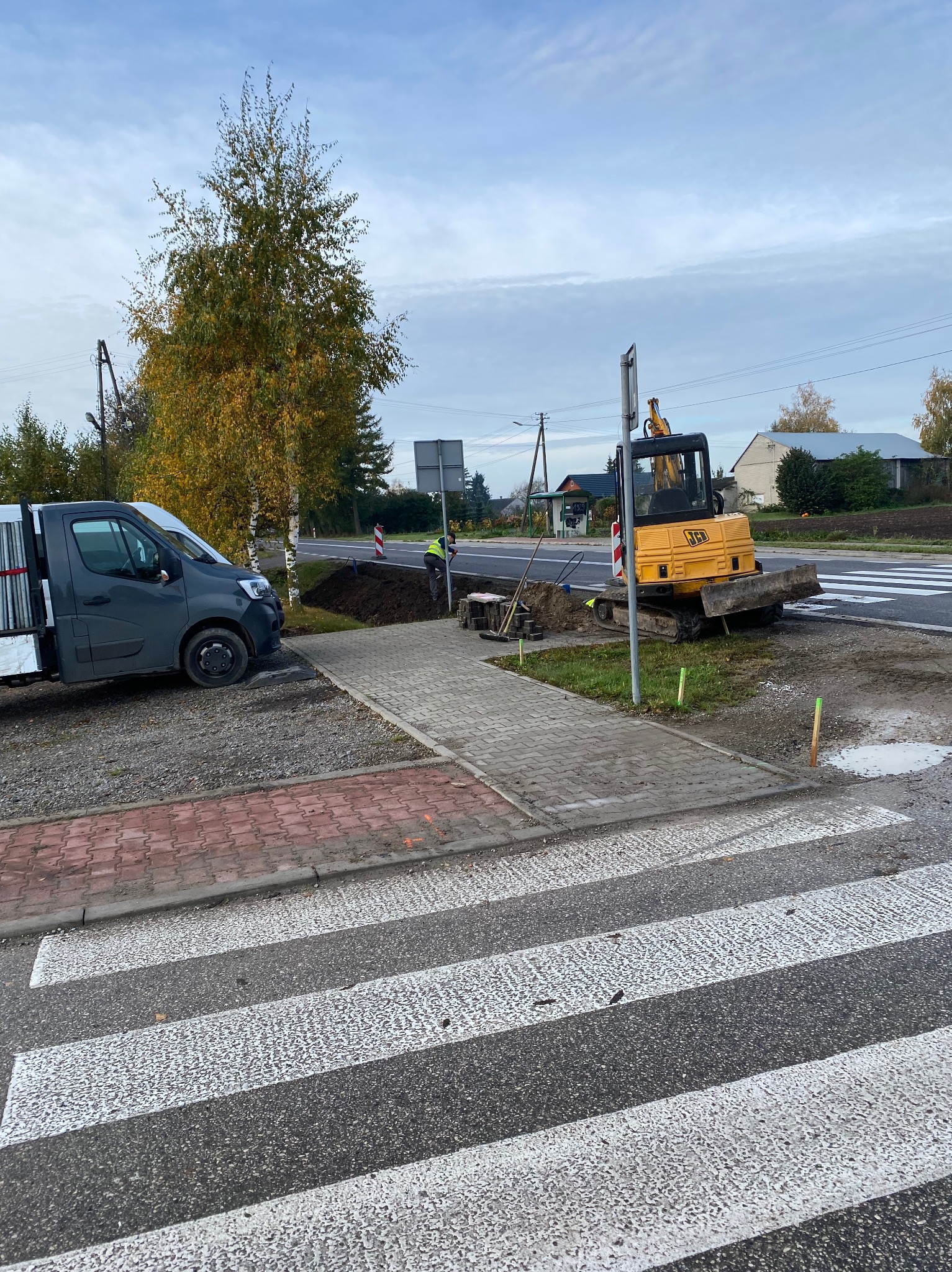 Żółta mini koparka JCB przy wykopie obok chodnika i przejścia dla pieszych, obok zaparkowany szary bus z rurami na pace, pracownik w kamizelce odblaskowej przy wykopie.