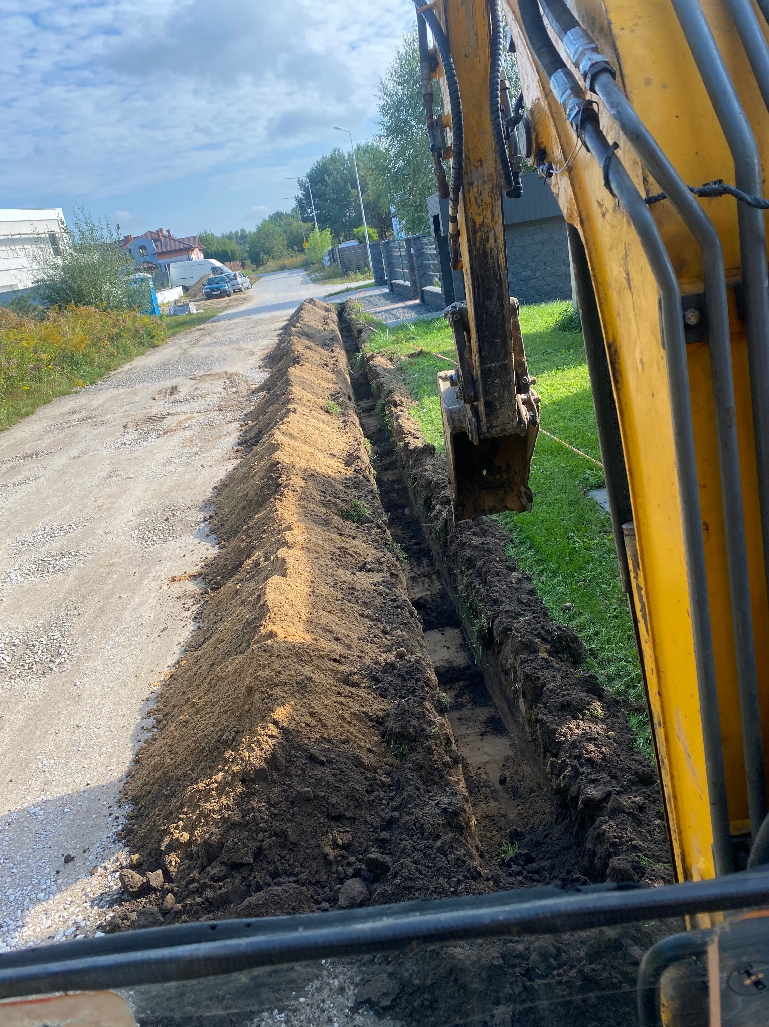 Żółta koparka wykopuje wąski rów wzdłuż drogi gruntowej na tle zabudowy jednorodzinnej i pochmurnego nieba.