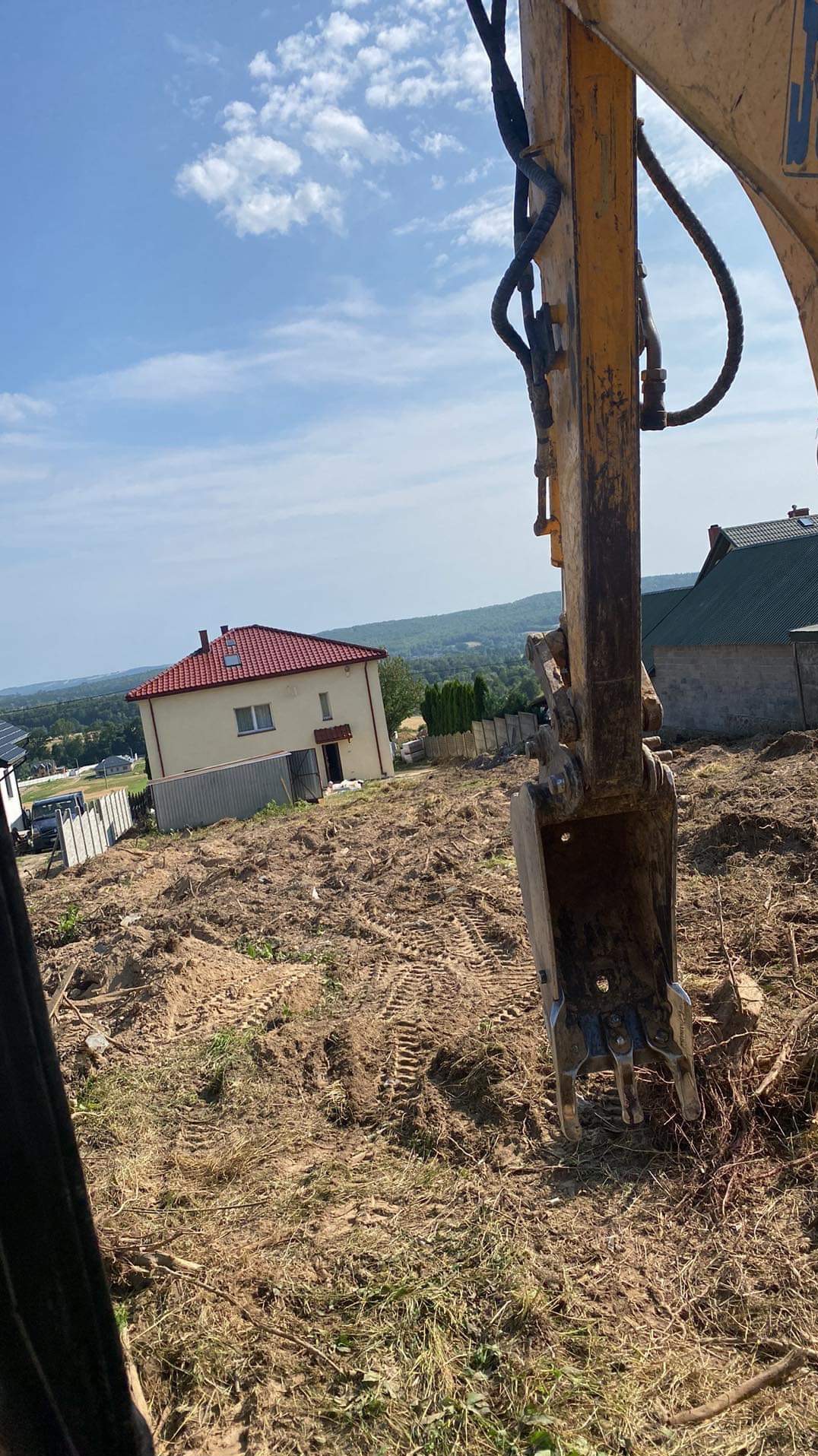 Żółta koparka z łyżką w trakcie niwelacji terenu pod budowę domu z czerwoną dachówką na wzgórzu, widoczne ślady gąsienic na ziemi.