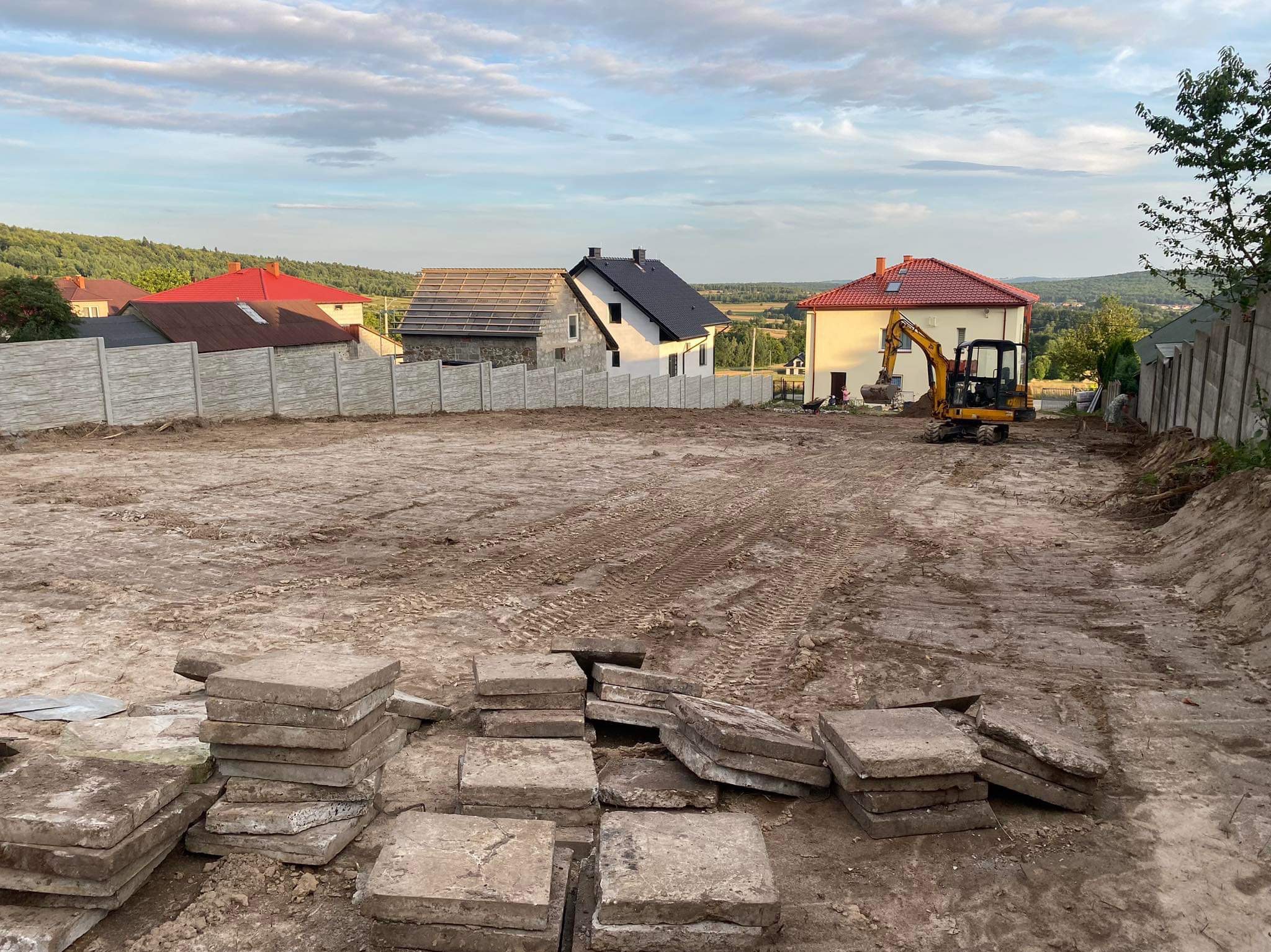 Żółta minikoparka na działce z przygotowanym terenem, w tle domy jednorodzinne i betonowe ogrodzenie, na pierwszym planie stosy betonowych płyt.