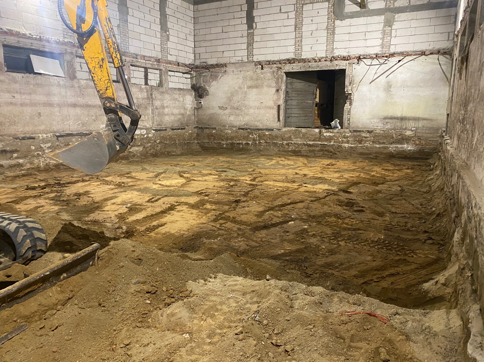 Żółta koparka w trakcie prac ziemnych wewnątrz budynku z surowymi ścianami z cegieł i betonu, widoczne ślady opon na wykopanej ziemi.