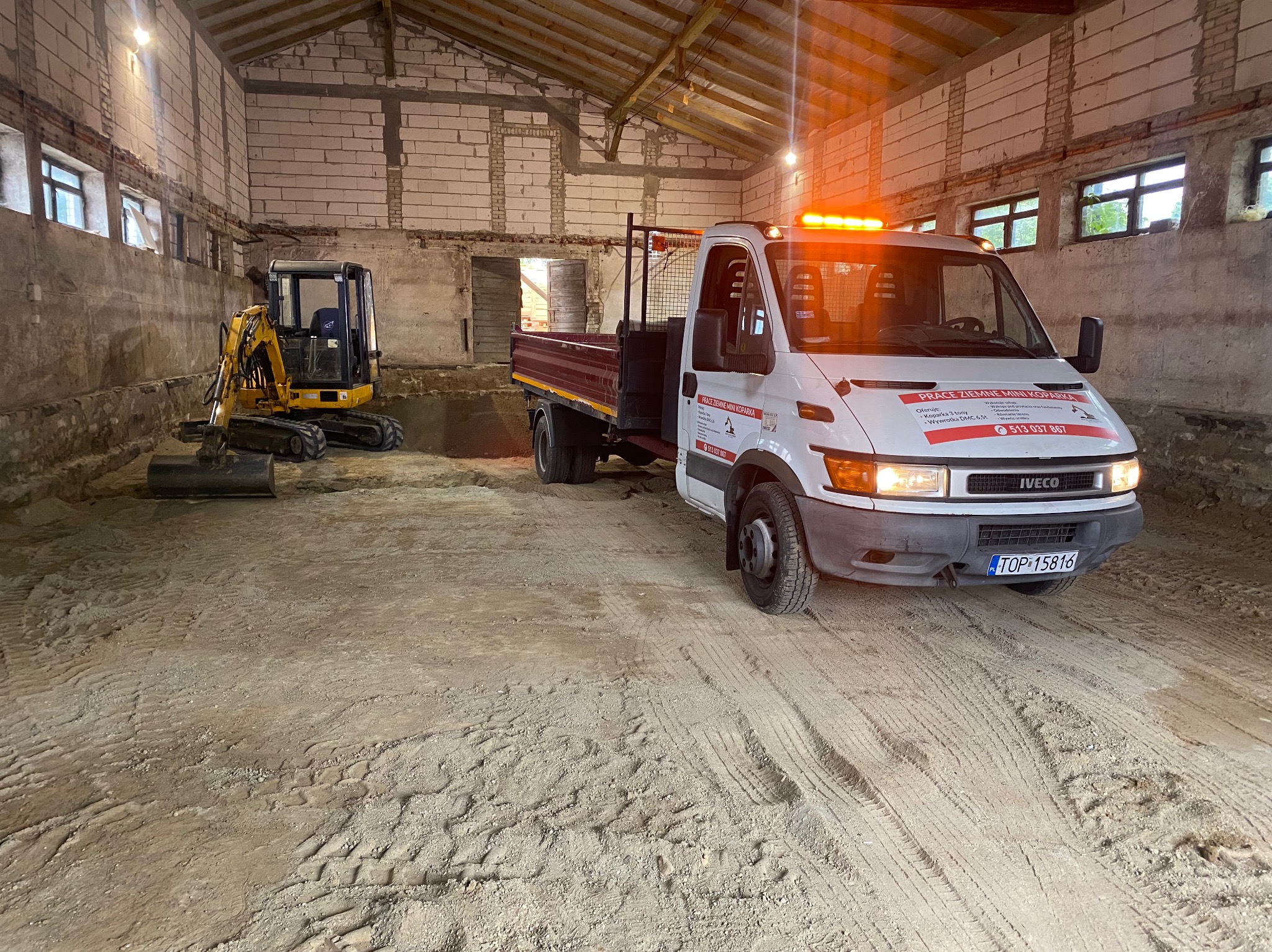 Wnętrze hali z mini koparką i wywrotką z pomarańczowymi światłami ostrzegawczymi, przygotowywane podłoże z widocznymi śladami opon i gąsienic.
