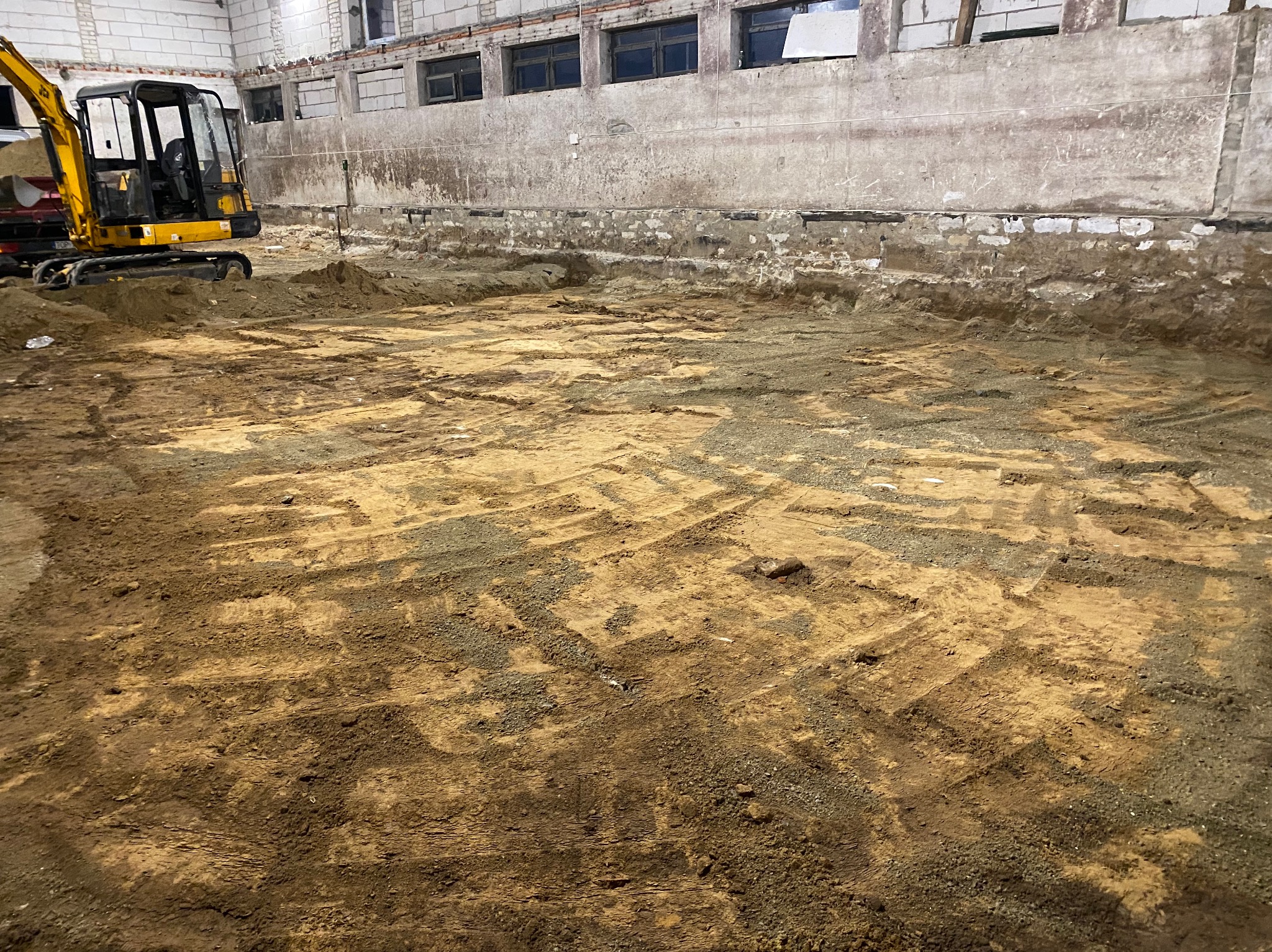 Wnętrze budynku w trakcie prac ziemnych, widoczny wykop z śladami opon i gąsienic oraz minikoparka w rogu, ściany betonowe z widocznymi uszkodzeniami i oknami pod sufitem.