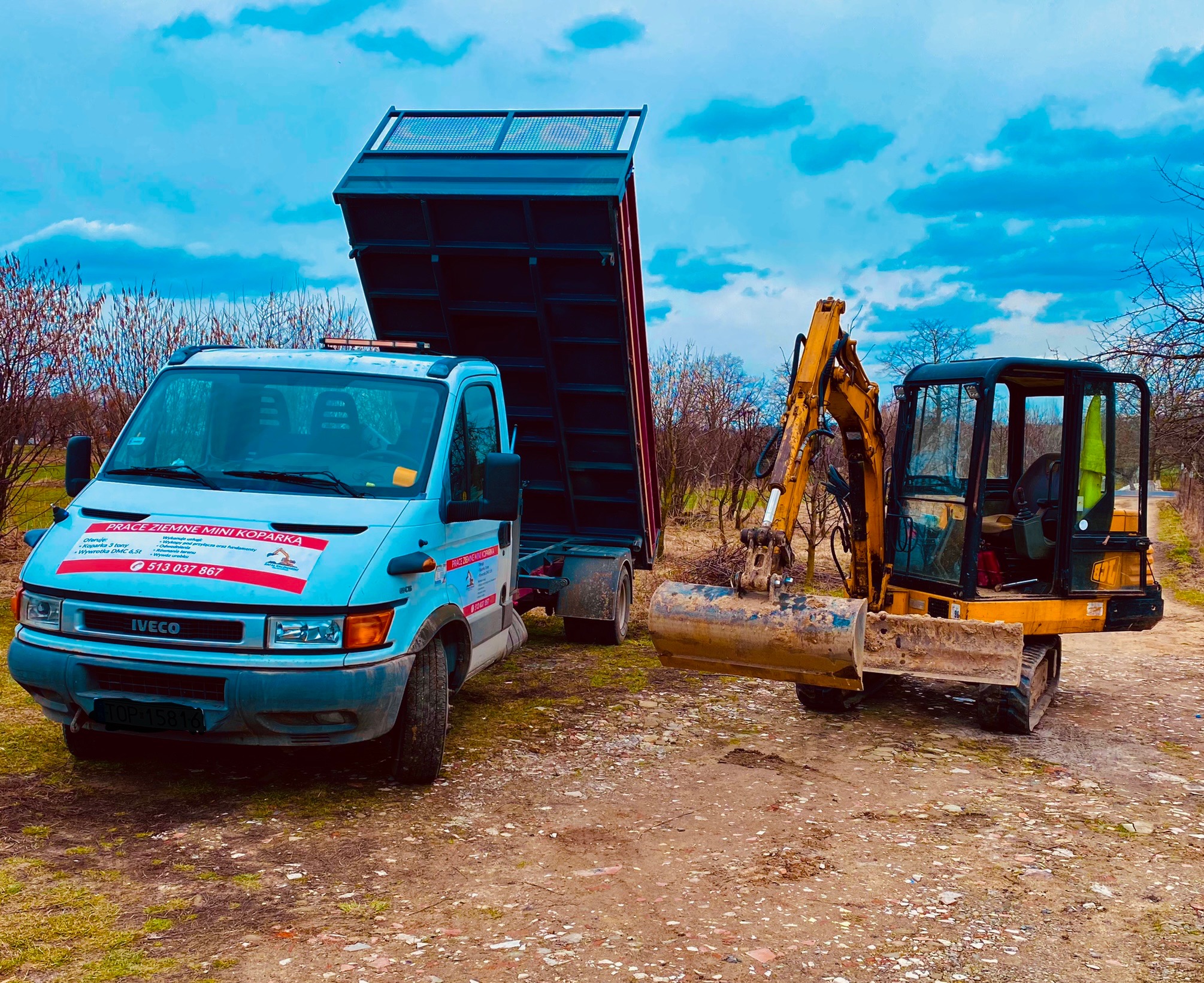 Żółta mini koparka z łyżką i Iveco z podniesioną skrzynią ładunkową na ziemnym podłożu, na pojeździe naklejka z ofertą prac ziemnych mini koparką.