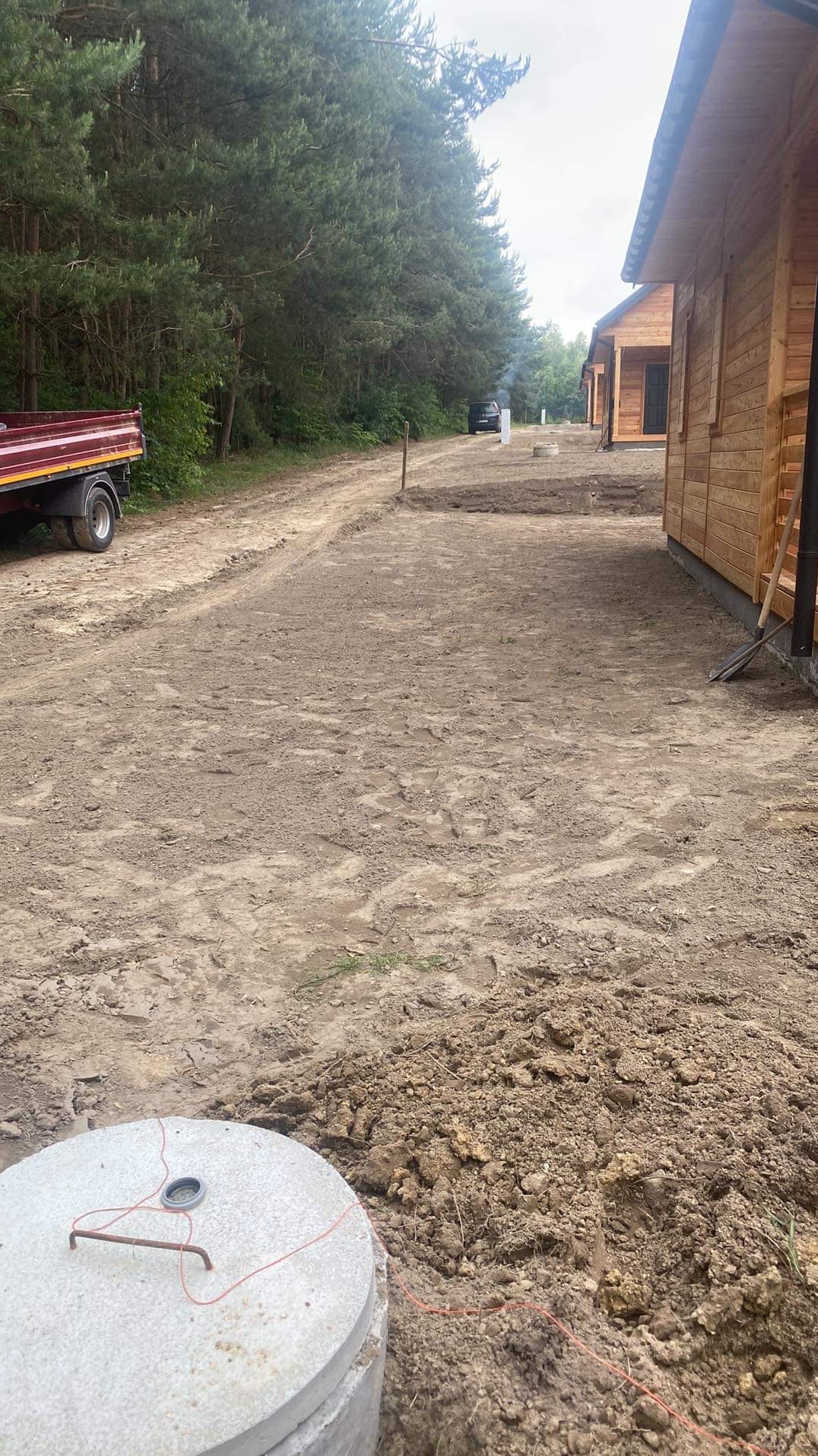 Przygotowany teren pod budowę domków letniskowych z widocznym wykopem pod szambo z betonowymi kręgami i wytyczonym obrysem, w tle las i samochód.