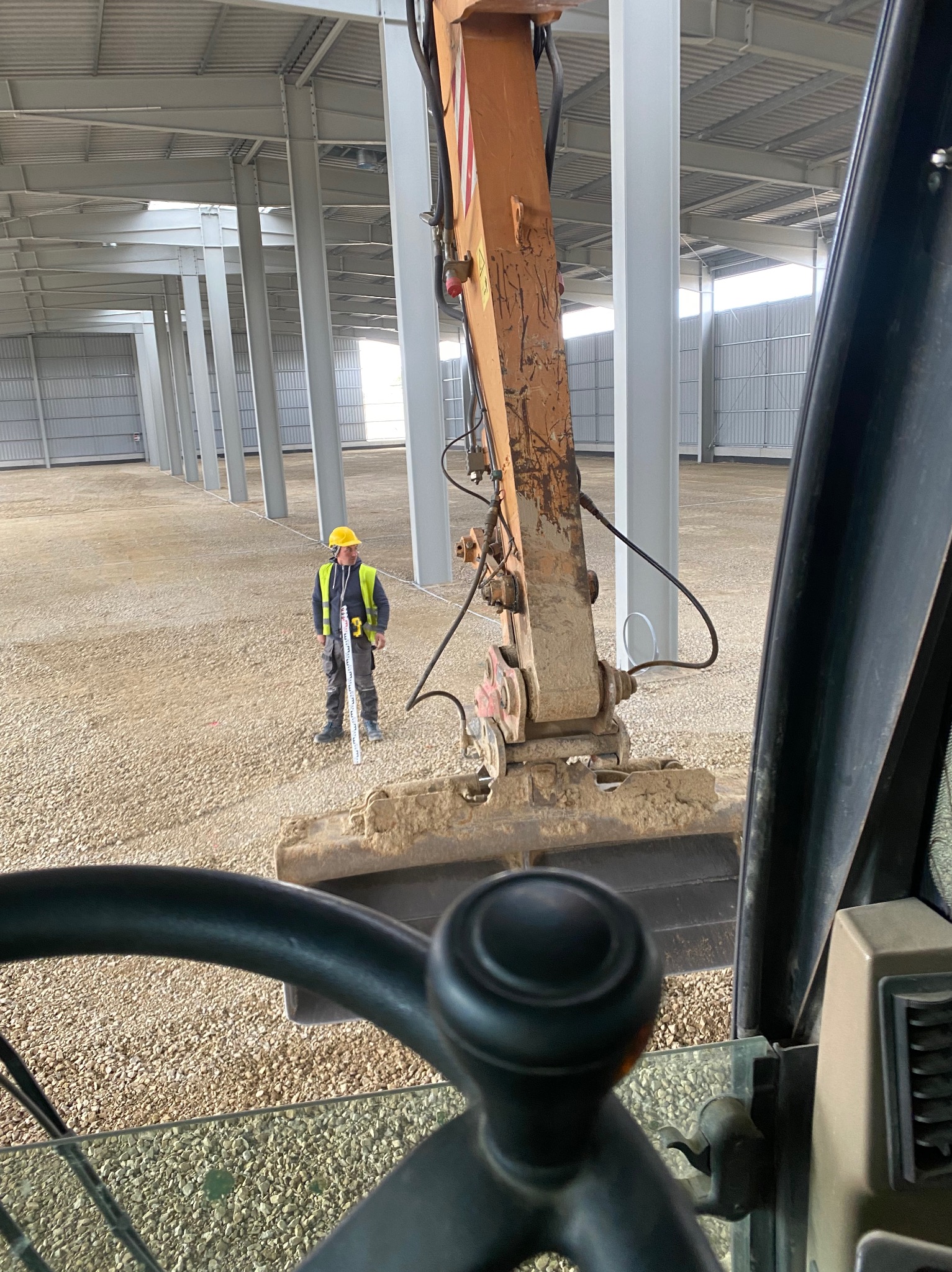 Widok z kabiny koparki na pracownika w żółtym kasku mierzącego poziom gruntu wewnątrz hali z metalową konstrukcją, przygotowanego pod budowę.