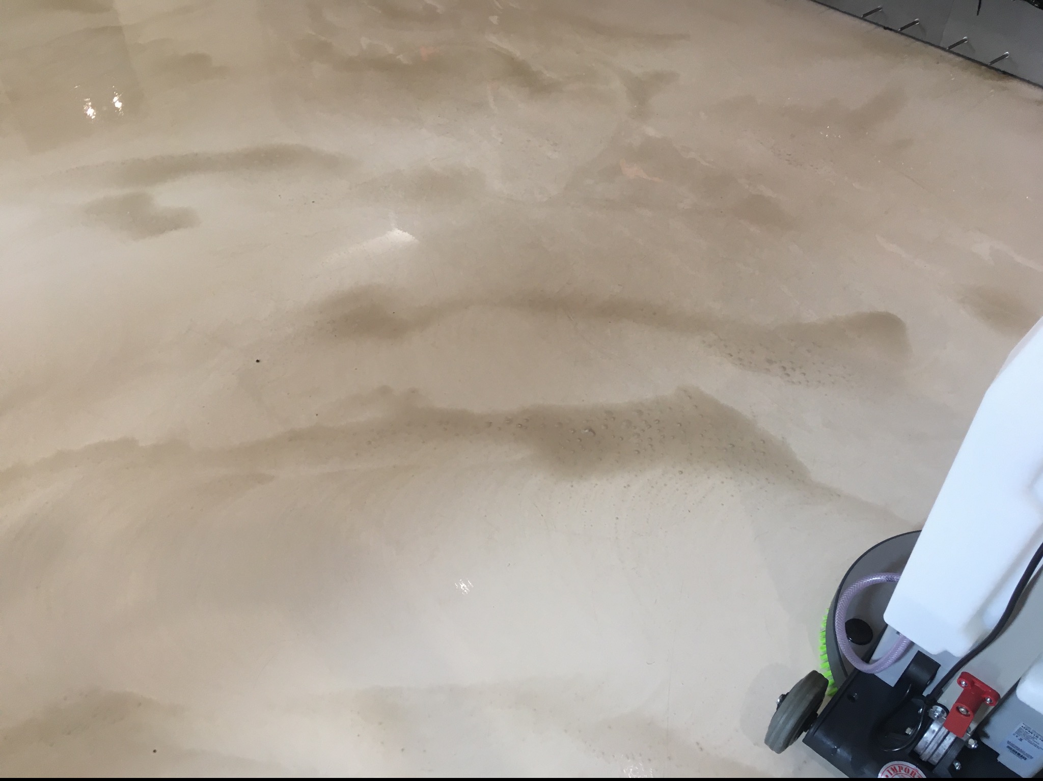 Automat szorujący w trakcie czyszczenia beżowej, błyszczącej podłogi z widocznymi smugami brudu i wody.