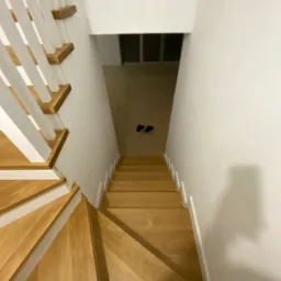 Widok z góry na drewniane schody z białymi poręczami w trakcie montażu, z widocznymi butami na podłodze w tle.