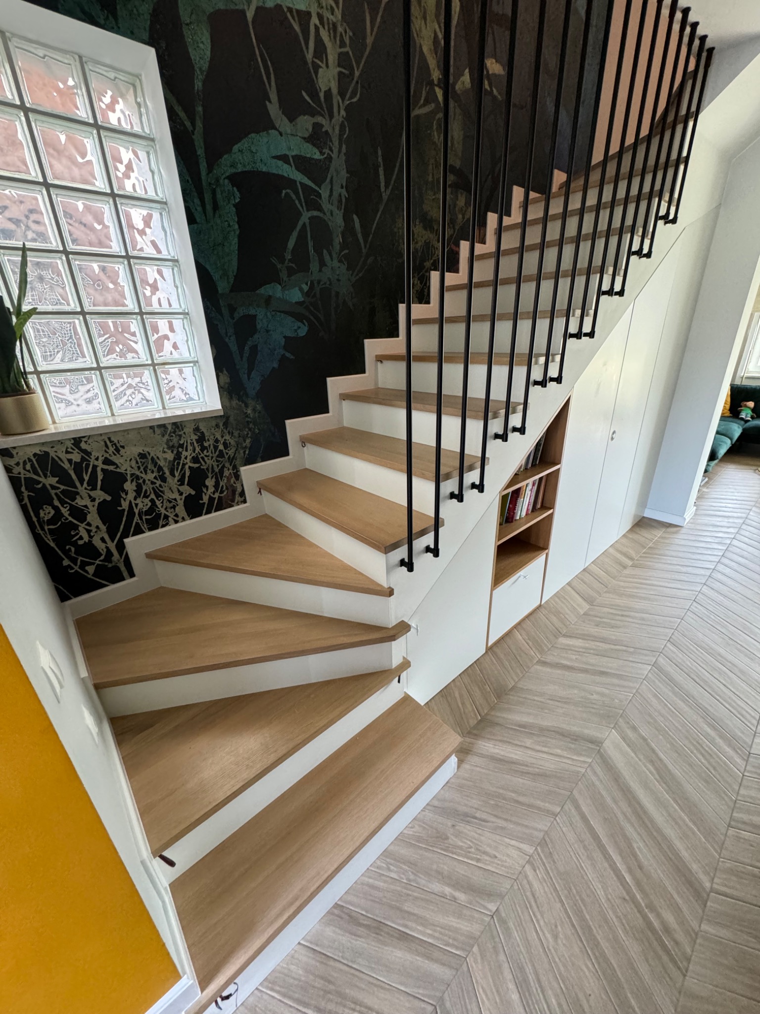 Bieg schodów z drewnianymi stopniami i czarną, minimalistyczną balustradą, zabudowany szafkami i regałem pod schodami, widok z góry na parkiet ułożony w jodełkę.