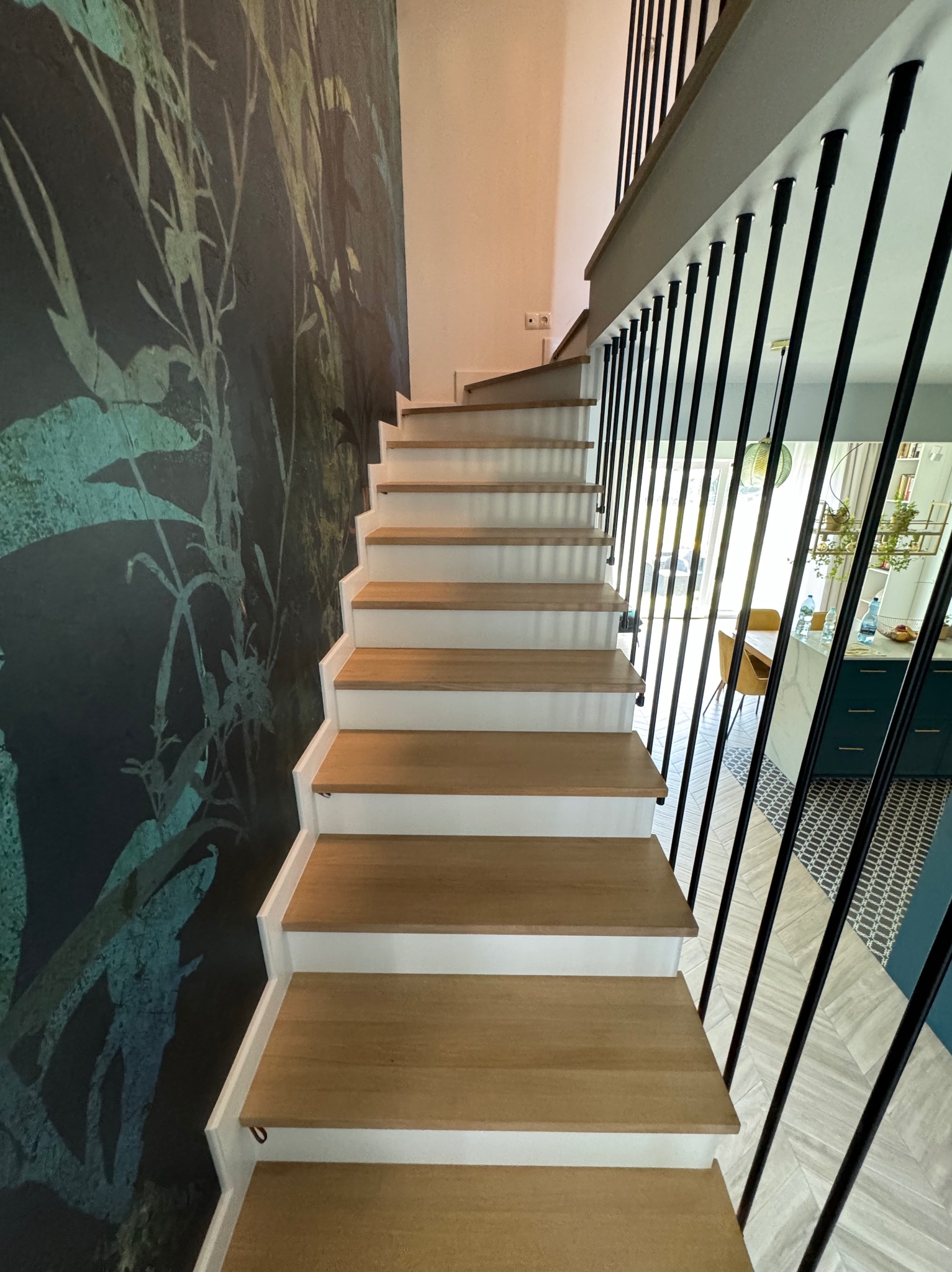 Nowoczesne schody drewniane z białymi podstopnicami i minimalistyczną, czarną poręczą w domu w Pułtusku; w tle widoczna ściana z motywem roślinnym i fragment kuchni.