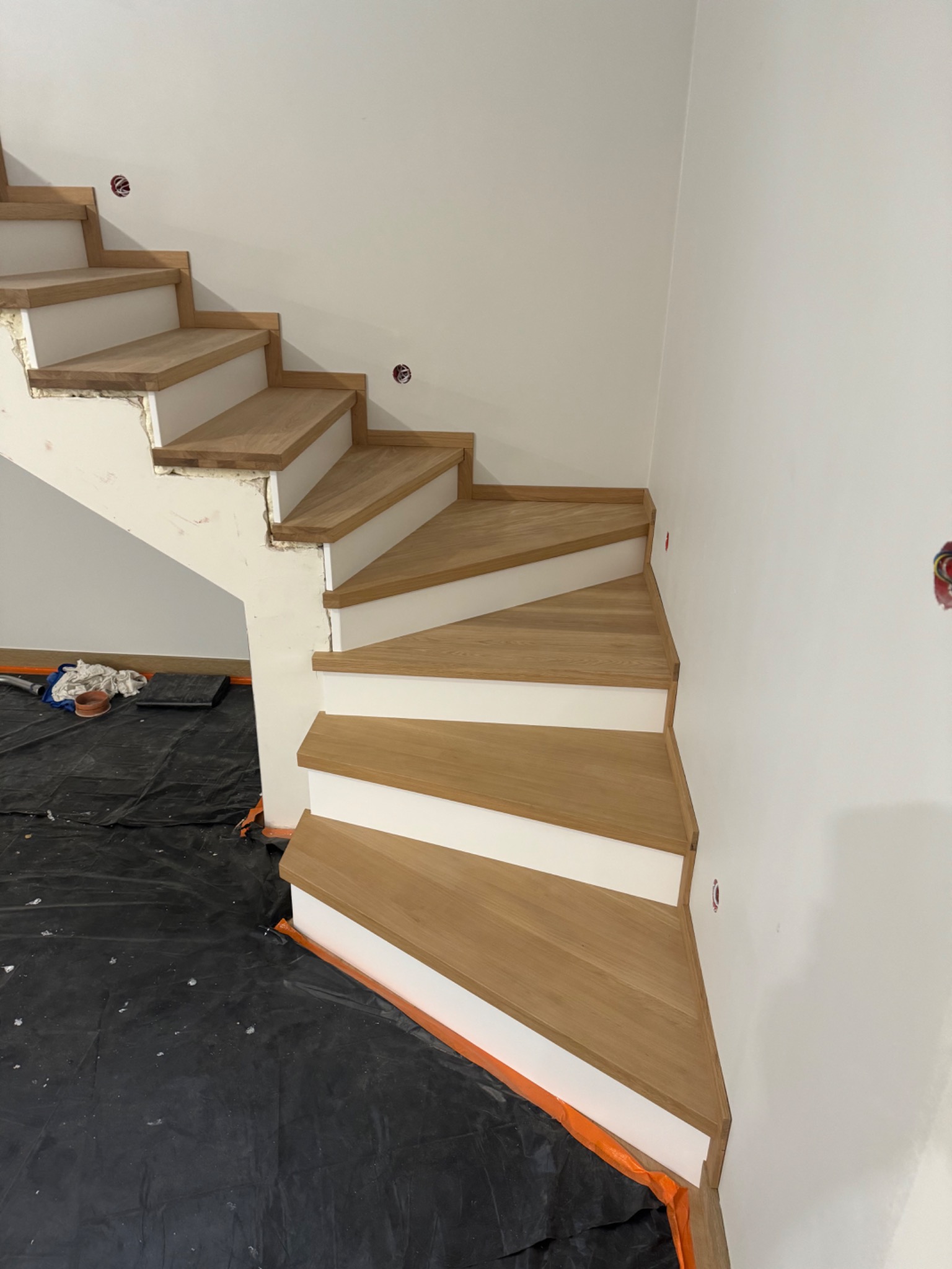 Montaż drewnianych schodów z białymi podstopnicami w trakcie wykańczania; widoczna czarna folia ochronna na podłodze i niezamaskowane puszki elektryczne w ścianie.