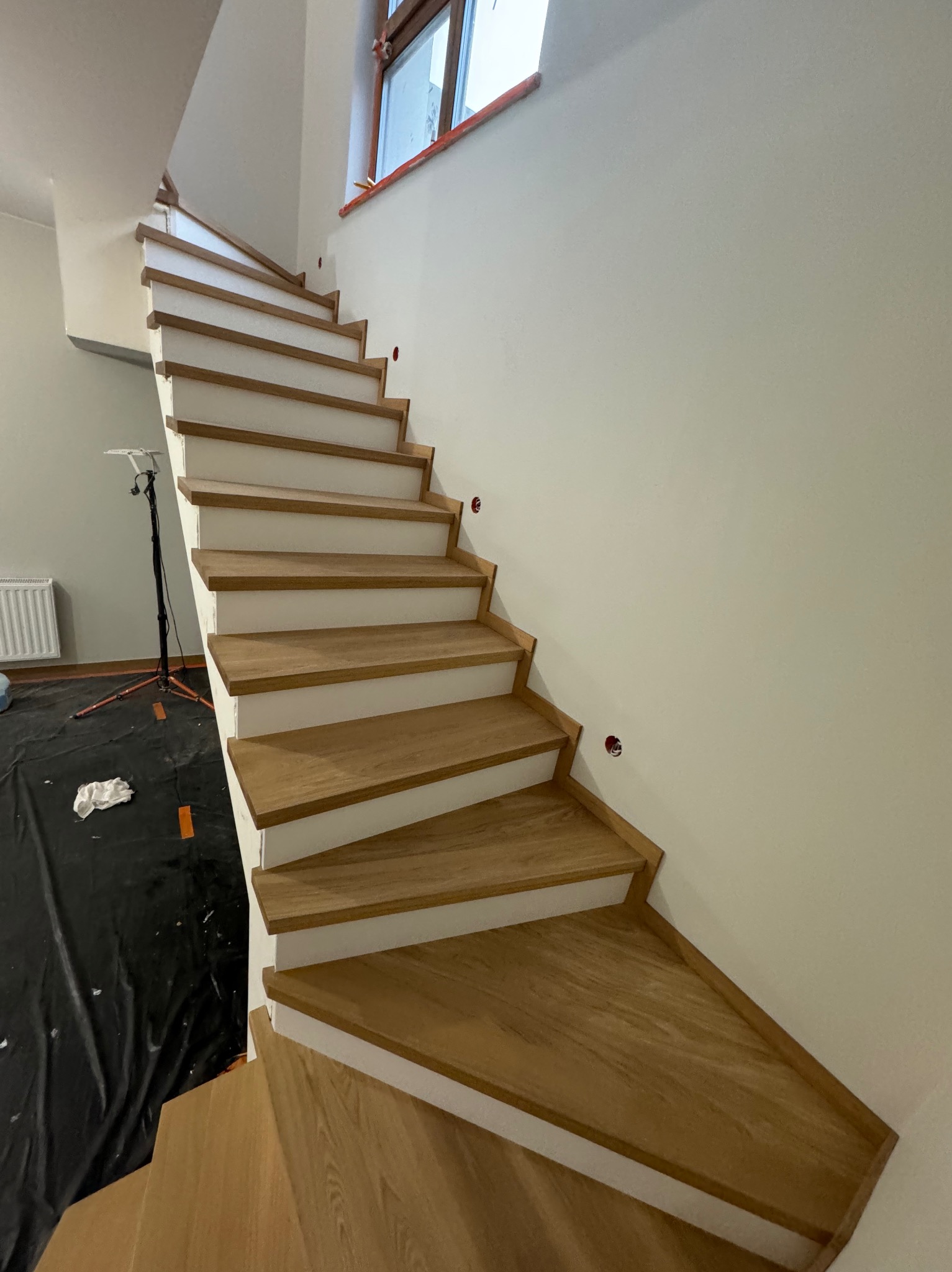 Wewnętrzne schody z drewnianymi stopniami i białymi podstopnicami, widoczne zabezpieczenia podłogi i lampa budowlana, sugerujące trwający montaż.
