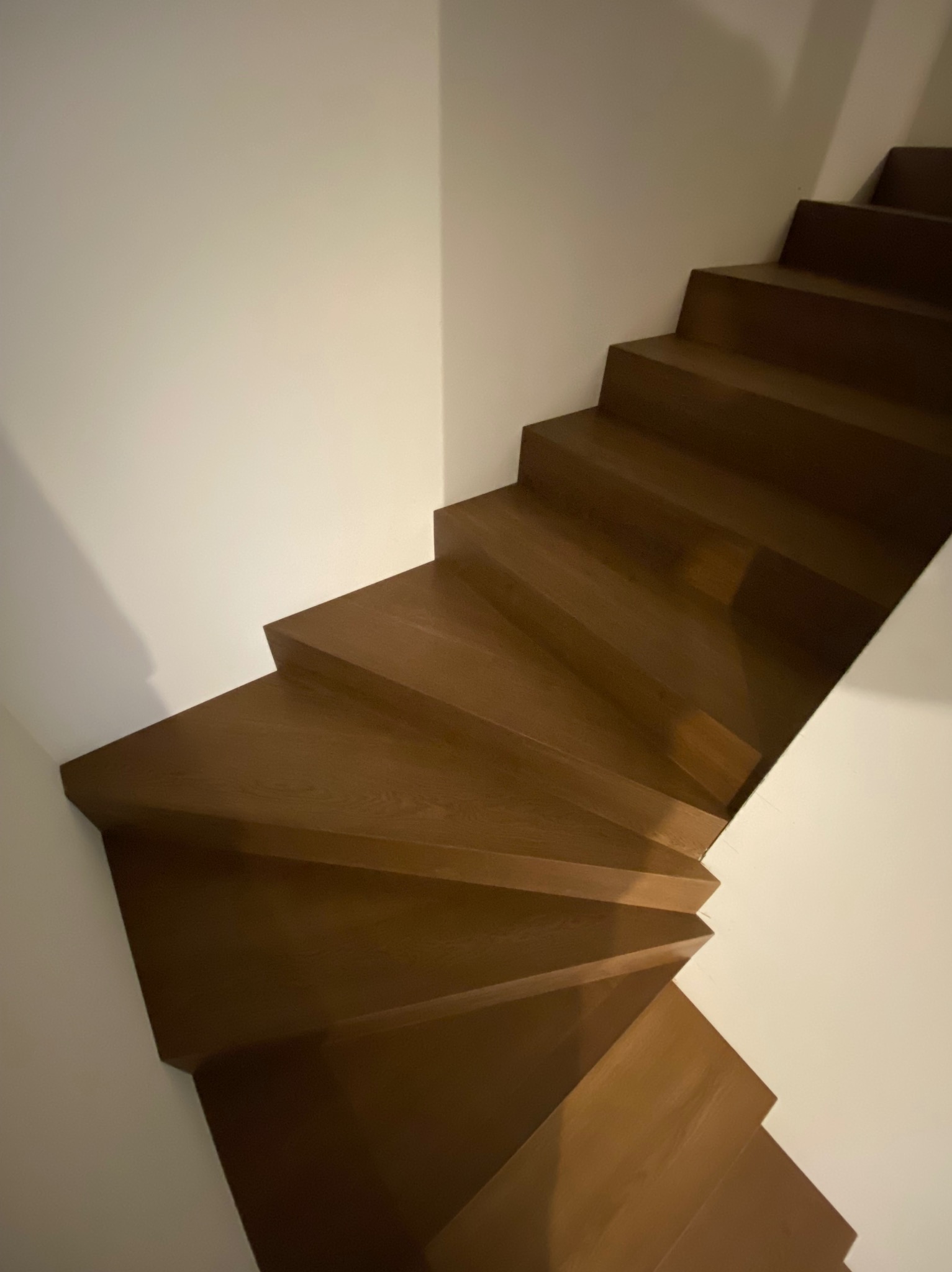 Kątowe ujęcie drewnianych schodów w kolorze orzecha, kontrastujących z białą ścianą, brak poręczy, widoczny zakręt schodów.