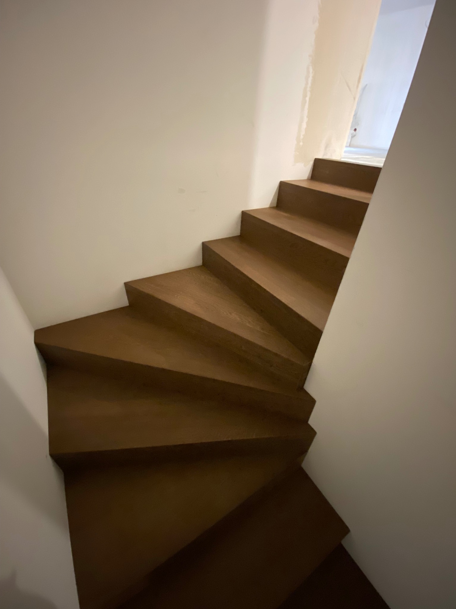 Wewnętrzne schody z drewna o ciepłym odcieniu brązu, skręcające w lewo, widoczne z dołu, z białymi ścianami w tle, częściowo wykończonymi.