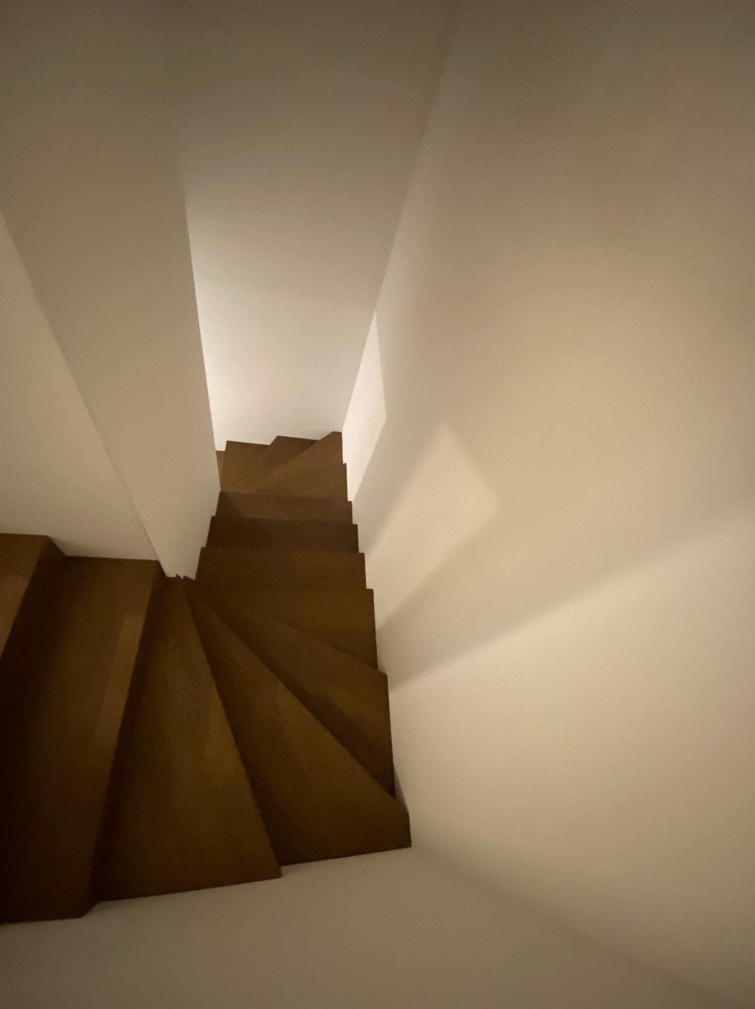 Wysoki kąt widzenia na drewniane schody zabiegowe z ciemnego drewna, prowadzące w dół, w otoczeniu białych ścian.