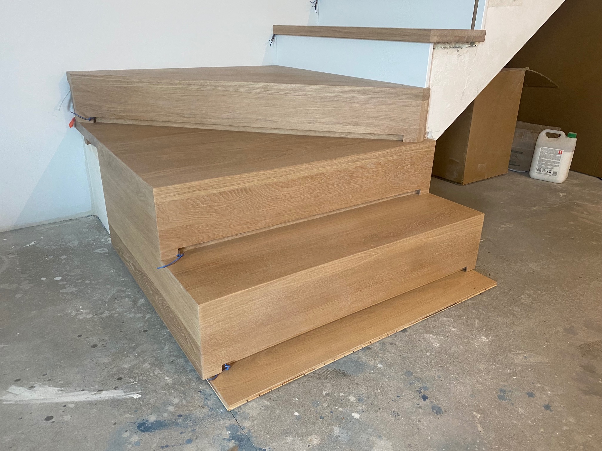 Surowe, dębowe stopnie schodowe w trakcie montażu, widoczne kable instalacyjne i elementy podkładowe na szarym, betonowym podłożu.