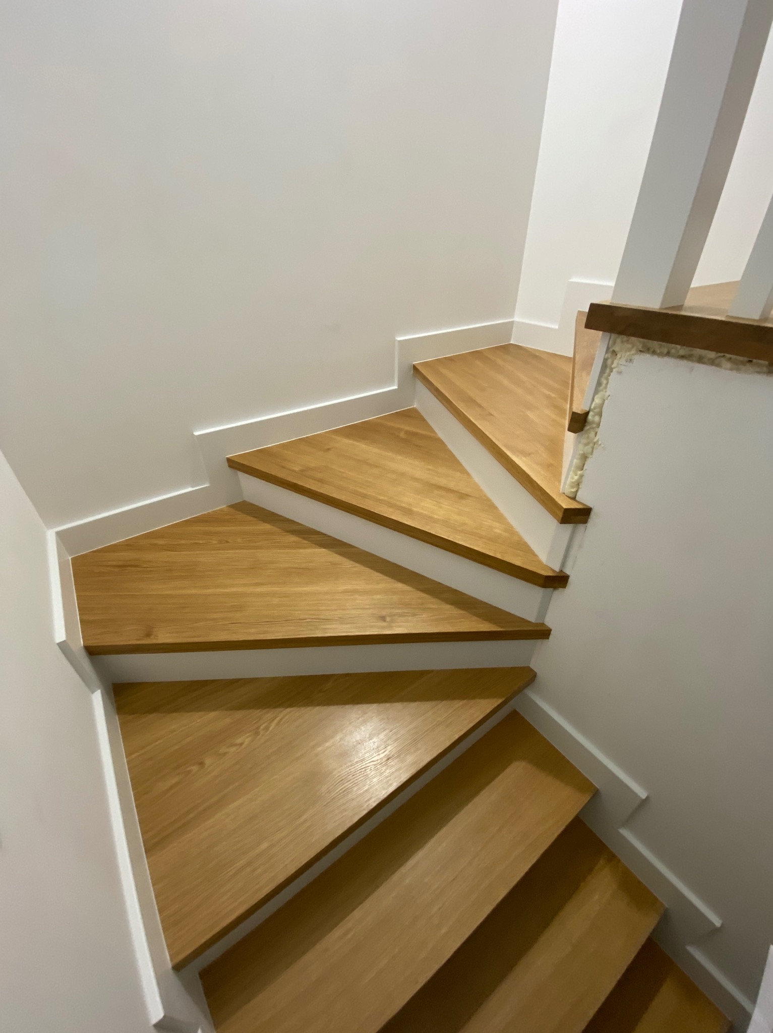 Drewniane schody w jasnym wnętrzu, widoczne z góry, z białymi listwami przypodłogowymi i fragmentem balustrady z pianką montażową.