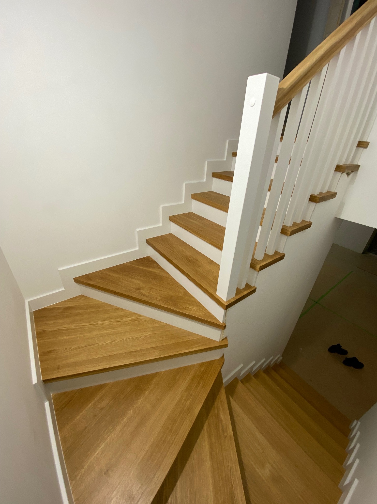 Widok z góry na drewniane schody z białymi podstopnicami i balustradą z drewnianą poręczą, widoczne buty na podłodze na dole.