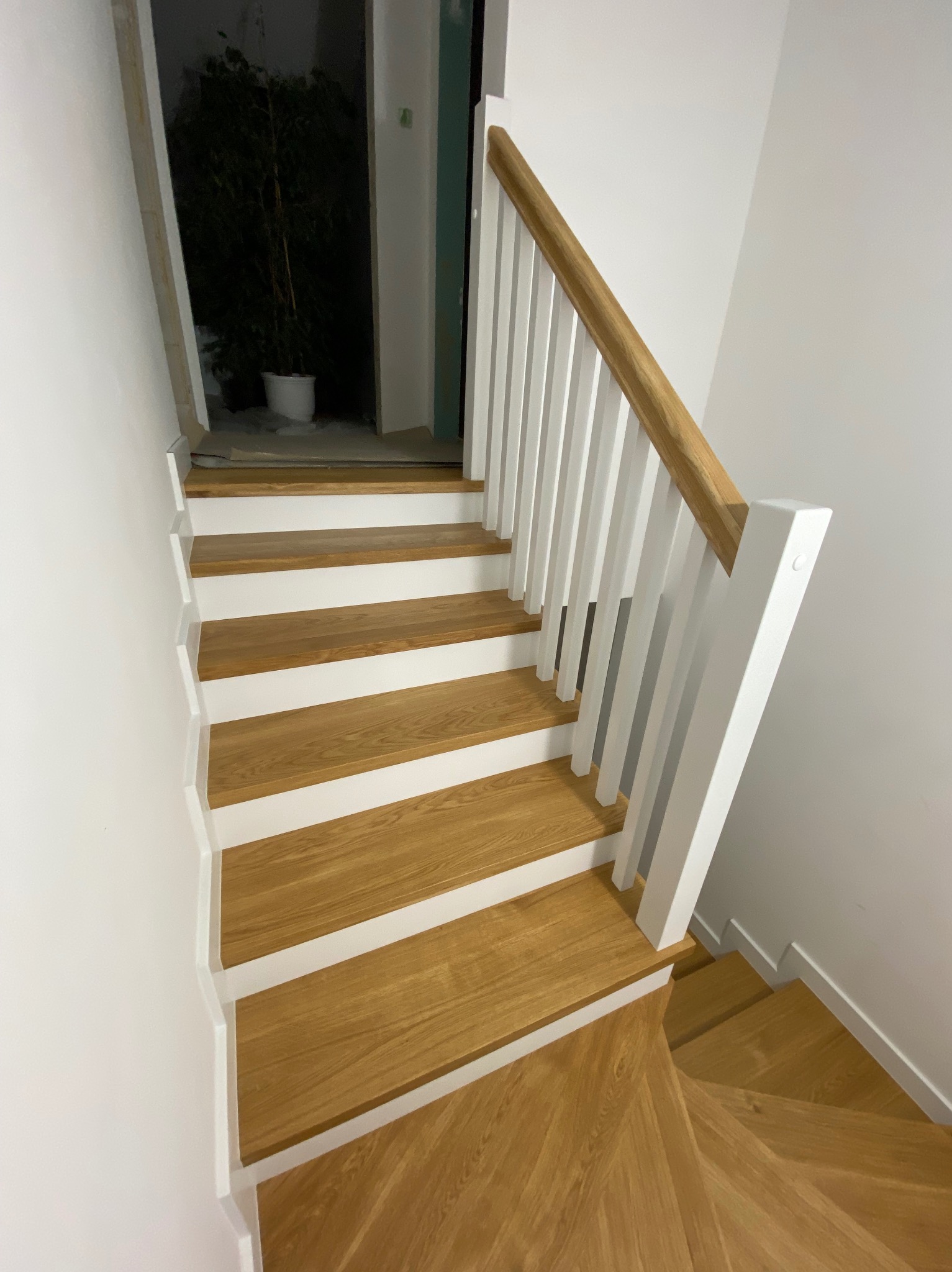 Jasne drewniane schody z białymi podstopnicami i poręczą z naturalnego drewna, widok z góry, w tle ciemne wejście z rośliną doniczkową.