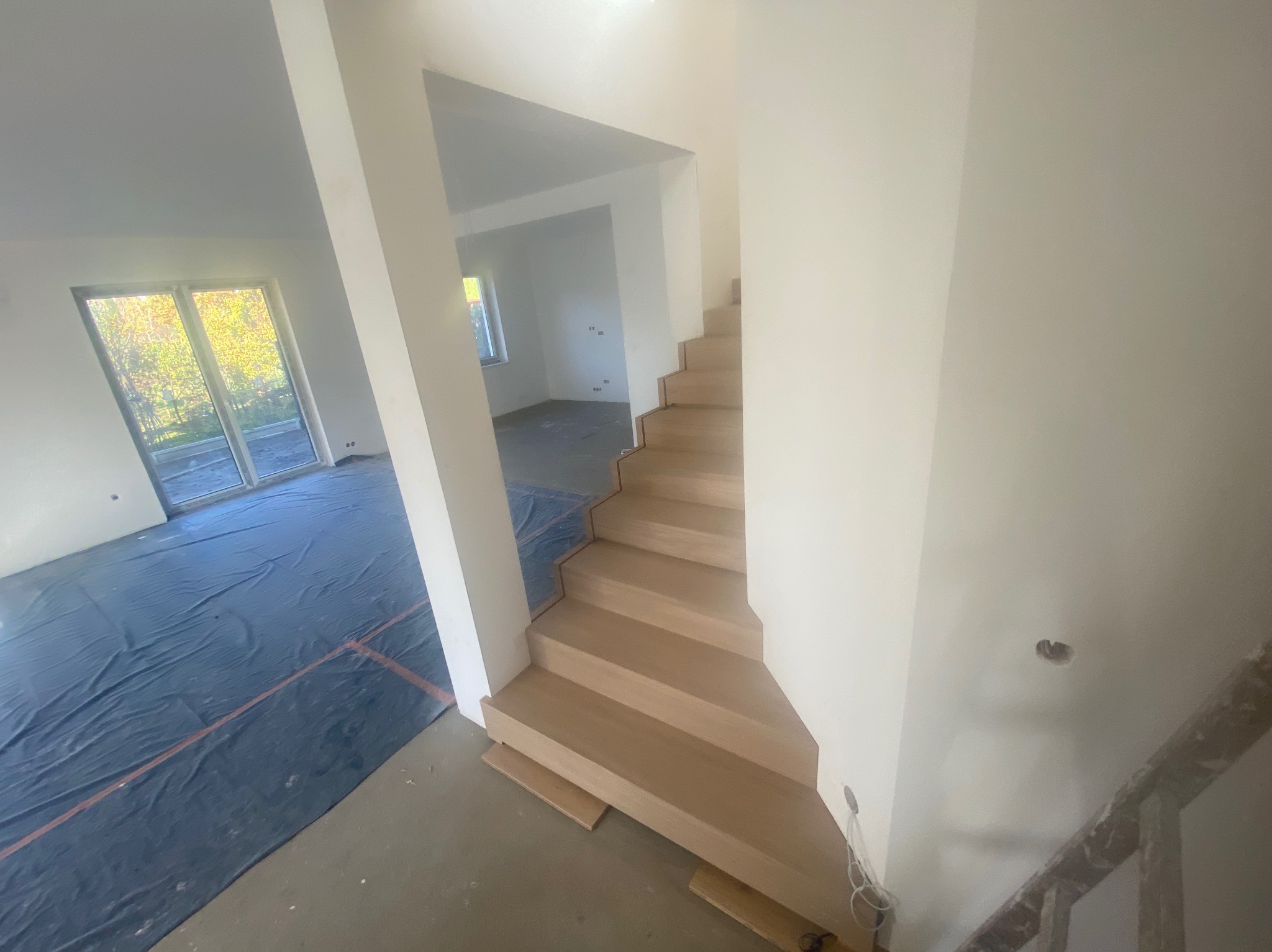 Jasne, drewniane schody bez poręczy w surowym wnętrzu domu w trakcie wykończenia, zabezpieczona podłoga folią ochronną.