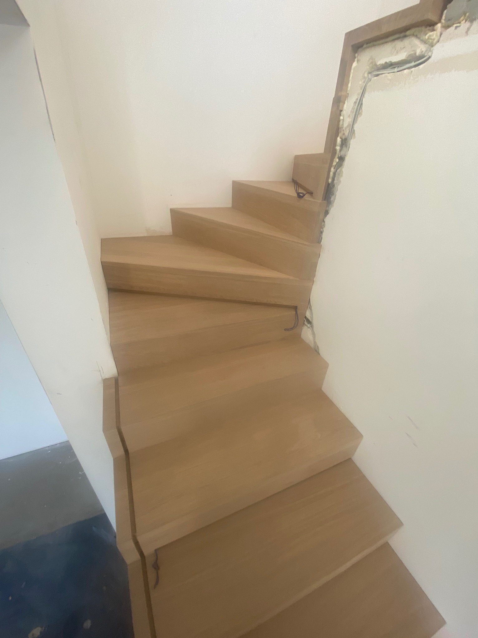 Widok z dołu na drewniane schody bez poręczy, w trakcie montażu, z widocznymi przewodami elektrycznymi w ścianie i surowym wykończeniem, oświetlone naturalnym światłem.