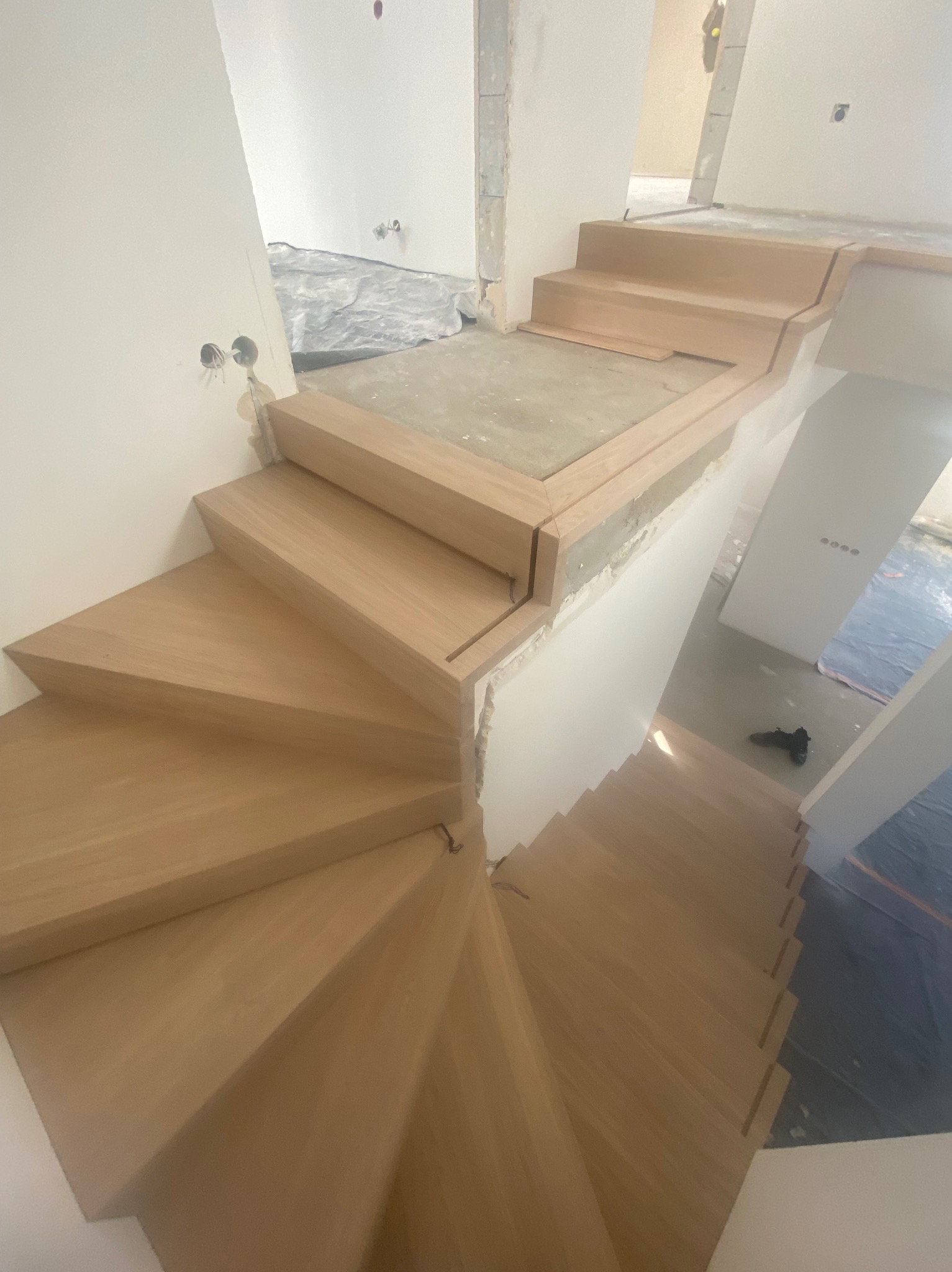 Wewnętrzne schody spiralne z jasnego drewna, w trakcie montażu, widoczne niedokończone ściany i zabezpieczenia podłogi.