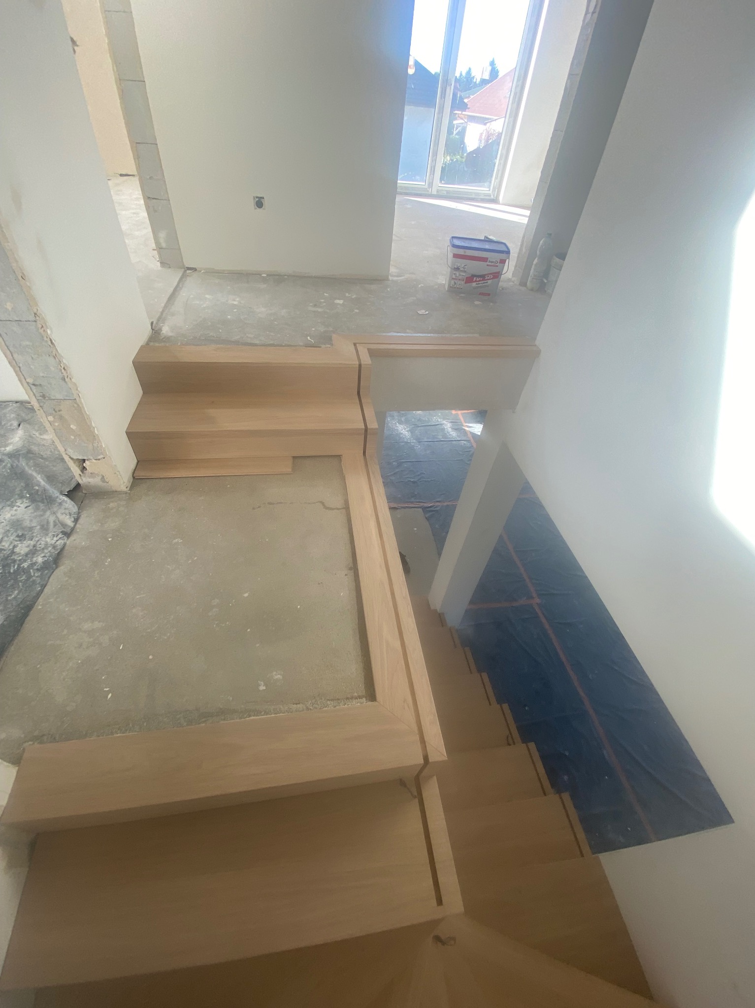 Montaż drewnianych schodów w surowym stanie w Pułtusku, widok z góry na pierwsze stopnie i zabezpieczone folią dalsze etapy prac.