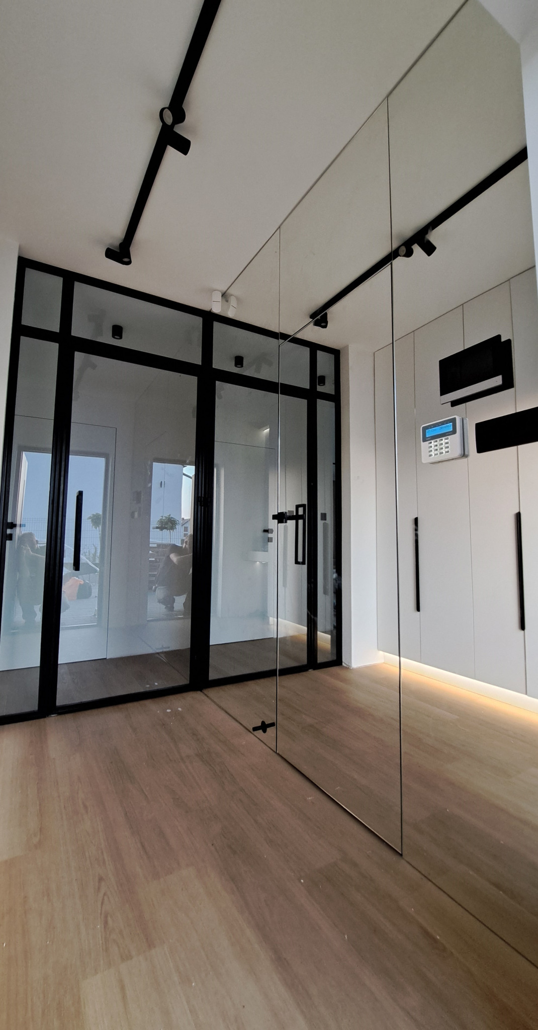 Nowoczesne wnętrze z przeszklonymi drzwiami w czarnych ramach, podświetlane panele ścienne i drewniana podłoga.