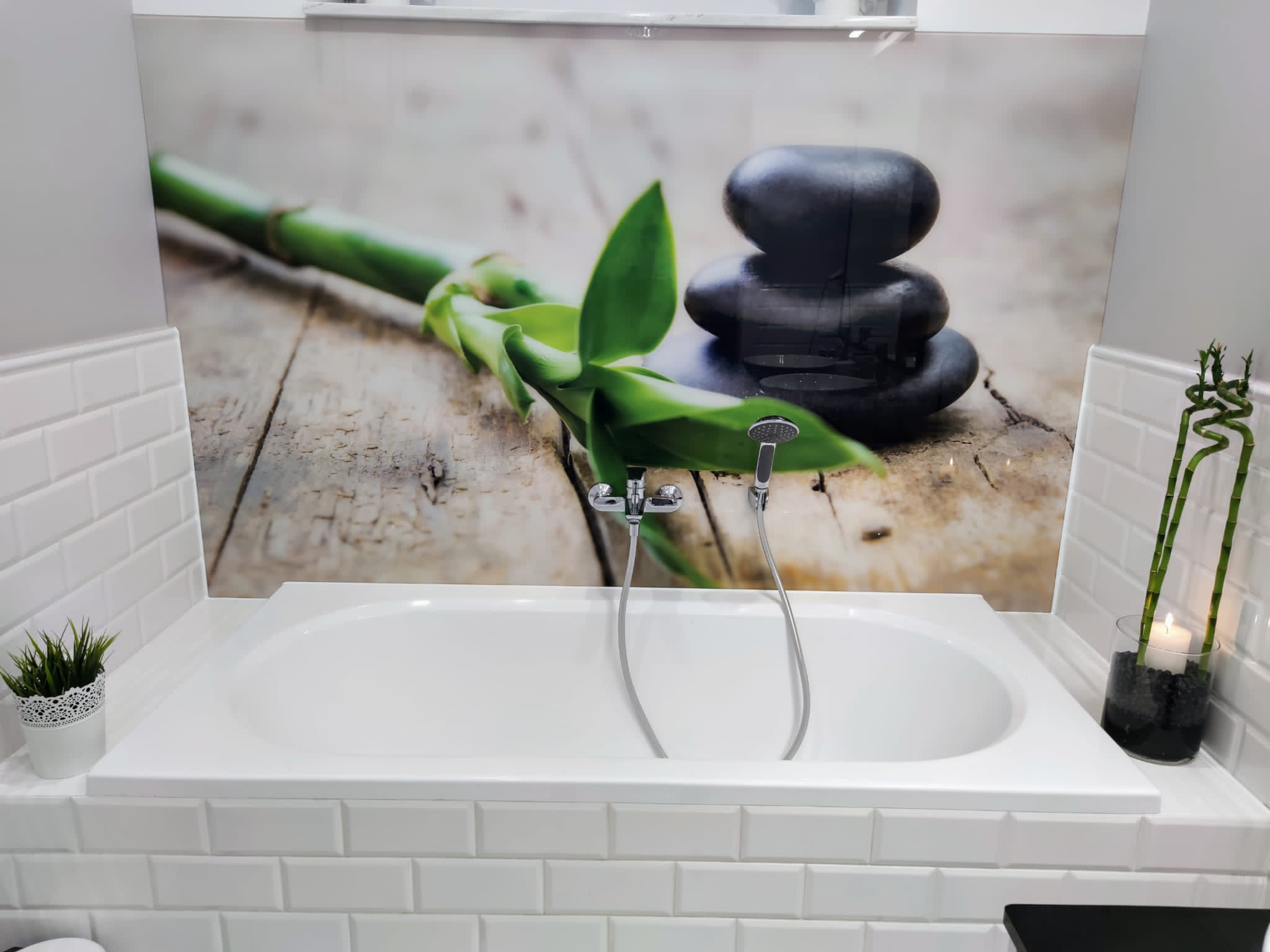 Biała wanna obudowana białymi płytkami, z baterią i słuchawką prysznicową, nad wanną panel dekoracyjny z motywem bambusa i czarnych kamieni, po bokach wazoniki z roślinami.